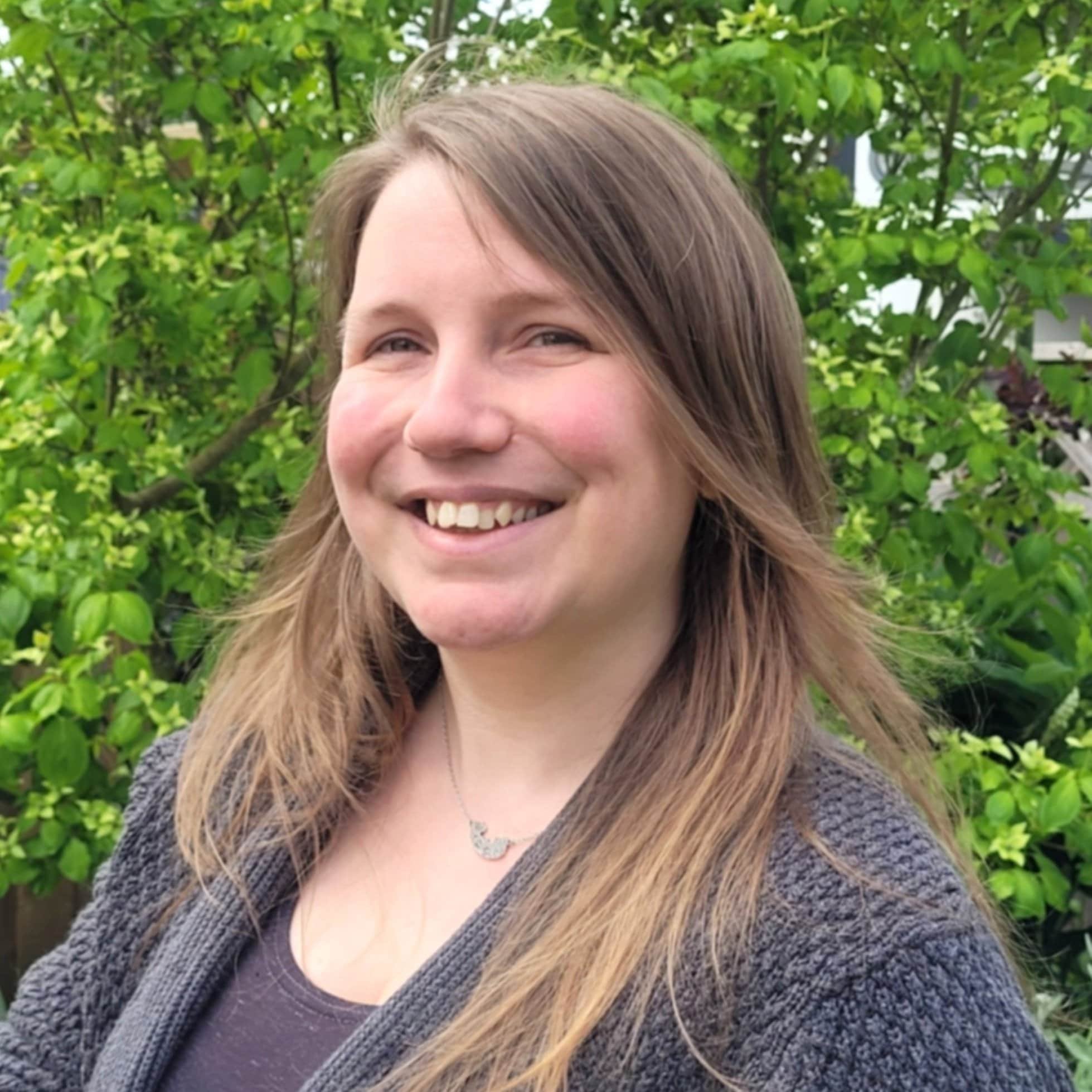 A smiling woman with long brown hair standing outdoors in front of green shrubbery.