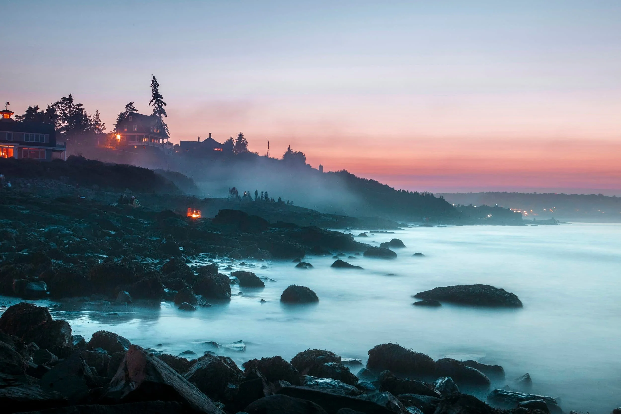 Rocky coastline at dusk with mist, glowing fires, and houses on a hill under a colorful pink and blue sunset sky.