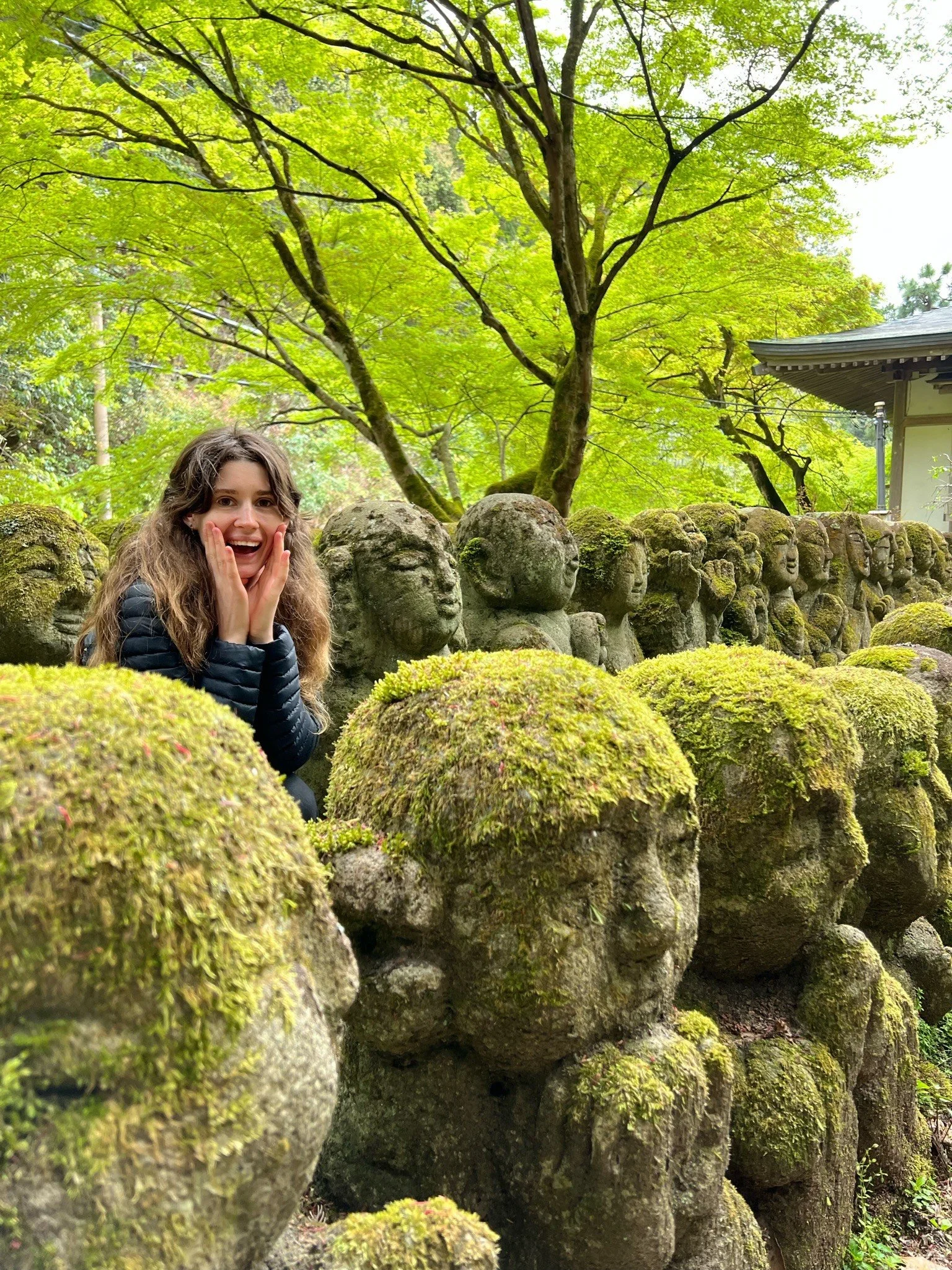 A woman with long curly hair, dressed in black, posing with surprise and joy in front of moss-covered stone statues in a lush green garden with trees and a small house.