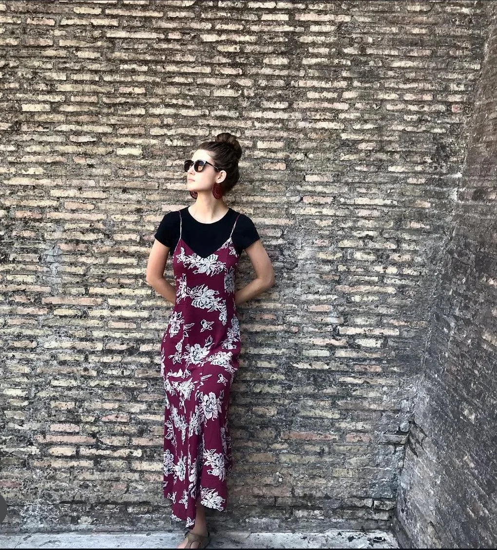 Woman wearing sunglasses and earrings, standing against a brick wall, dressed in a black t-shirt layered with a maroon floral slip dress. She has her hair up in a bun.