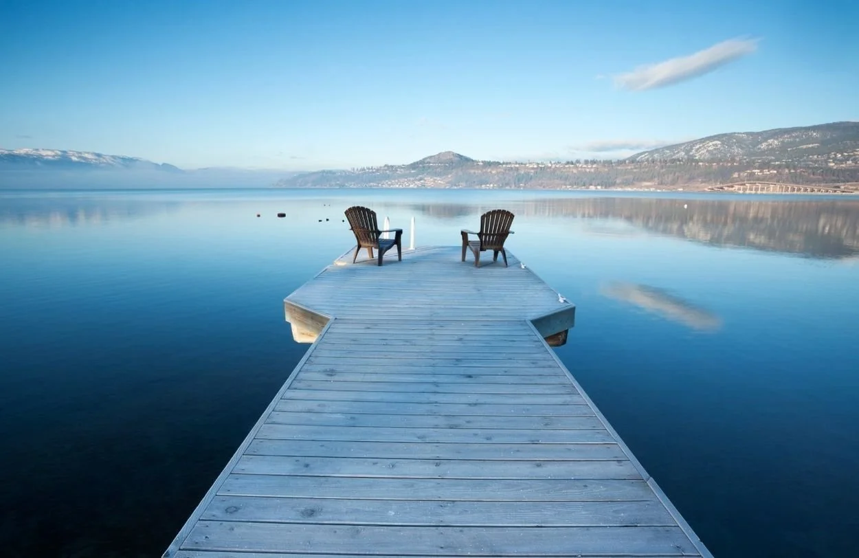A wooden dock extending into a calm, reflective body of water with two Adirondack chairs at the end, facing a scenic landscape of distant mountains and a blue sky with scattered clouds. Setting is BC where we do relationship therapy and sex therapy
