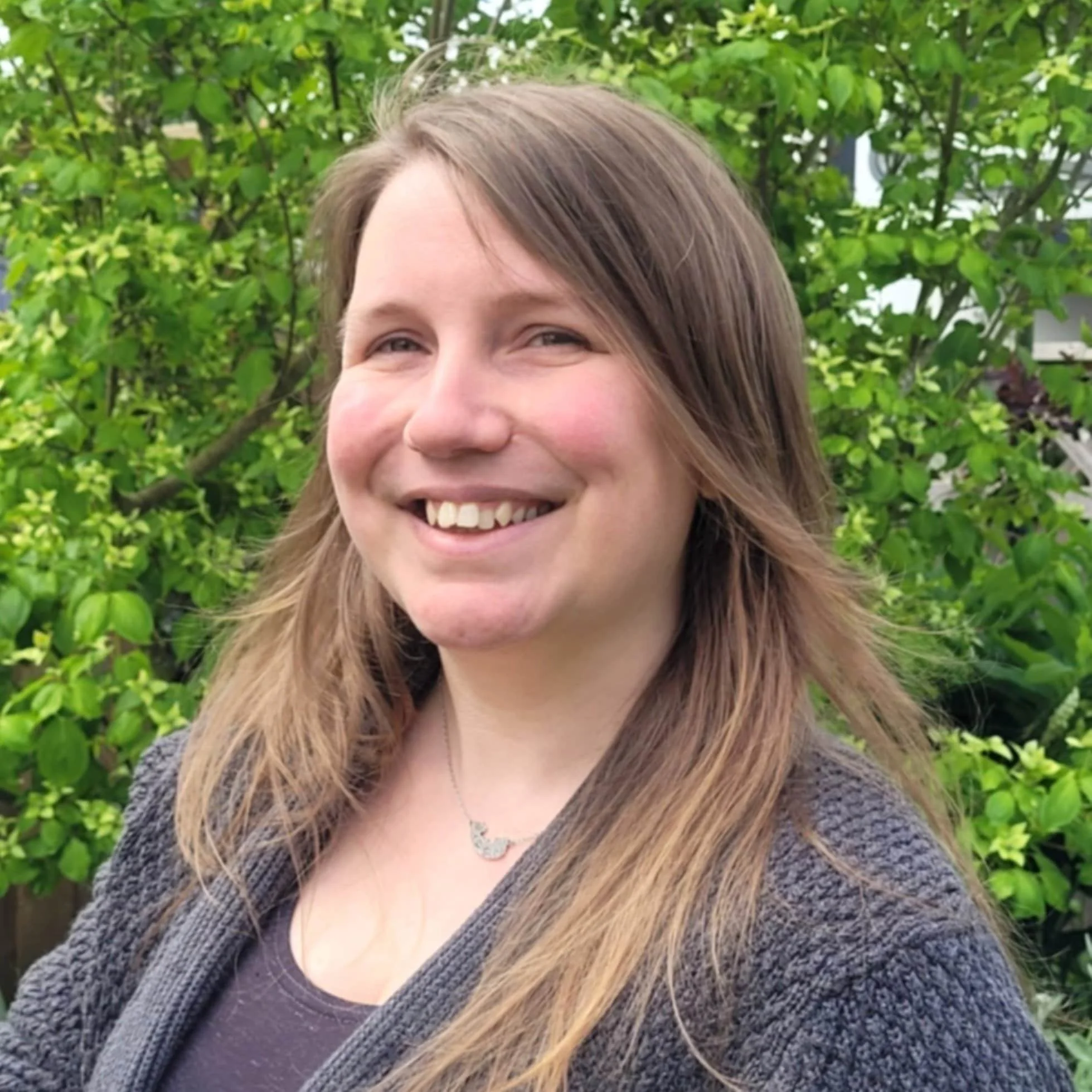 A smiling woman with long brown hair standing outdoors in front of green foliage, wearing a dark T-shirt, a gray cardigan, and a silver necklace.