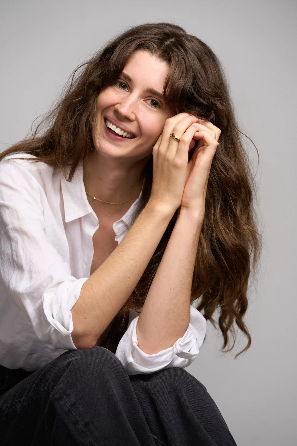 Close-up of a woman with long, wavy brown hair and green eyes smiling outdoors with green foliage in the background.