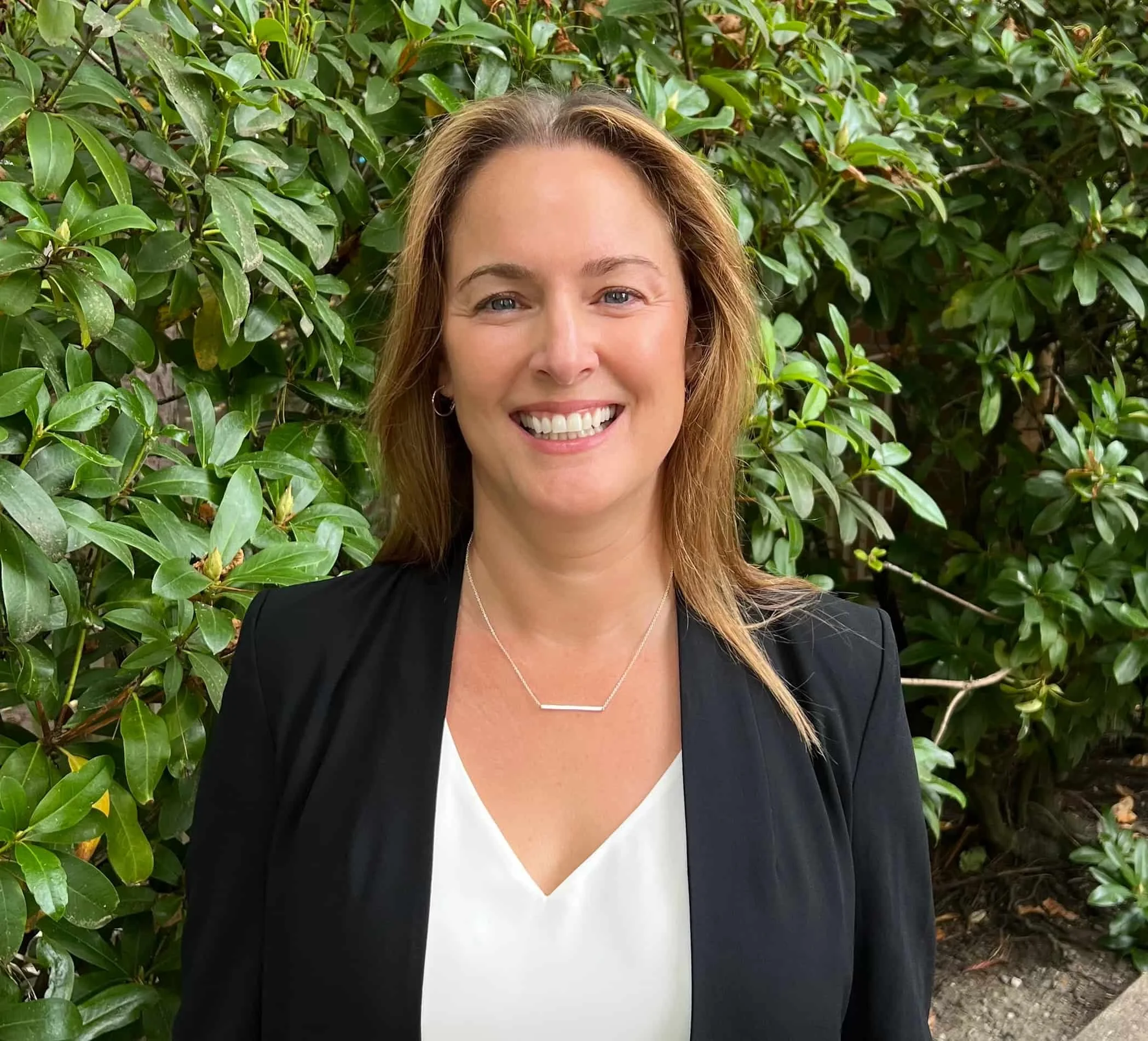 A woman with shoulder-length light brown hair, smiling, wearing a black blazer over a white blouse, standing outdoors in front of green leafy bushes.