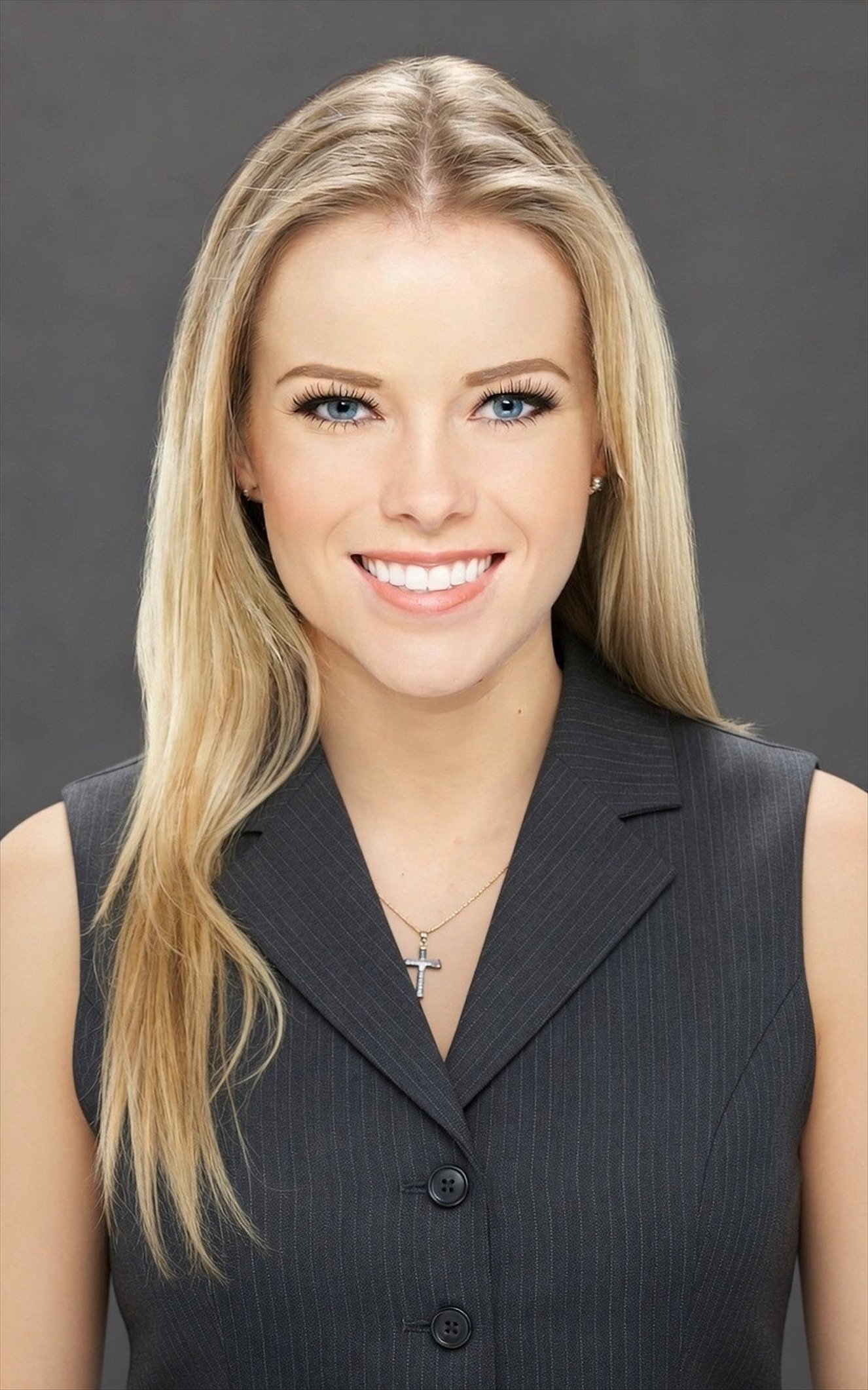 Portrait of a smiling woman with long blonde hair, wearing a blue collared shirt with a company logo. She has bright blue eyes and is set against a gray background.