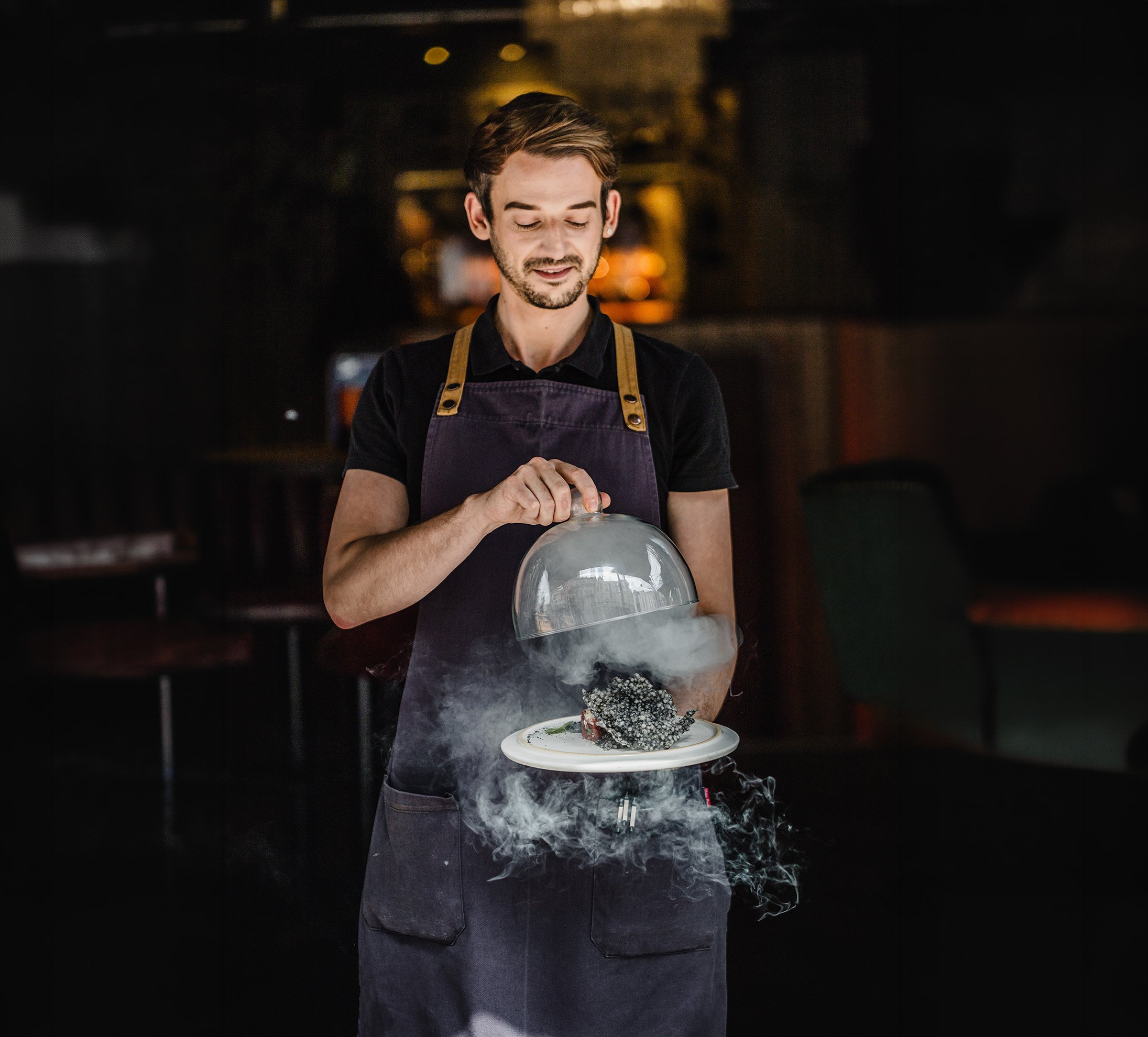 Environmental hospitality portrait of a smiling server presenting a plated dish beneath a raised glass cloche filled with smoke, capturing the personality and theatre of restaurant service for Casa Moa Bristol.