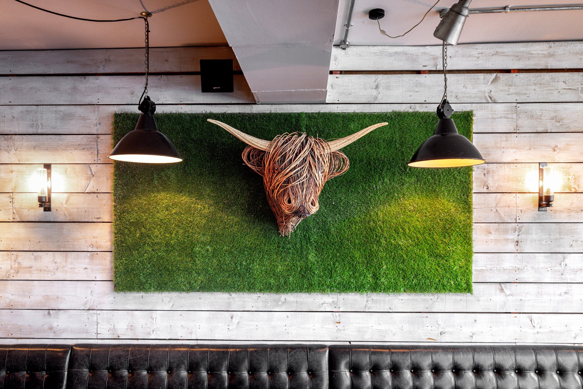 Interior design photograph at Pasture Restaurant in Bristol, showing a sculptural Highland cow head on a grass-covered wall framed by black pendant lights and pale timber.