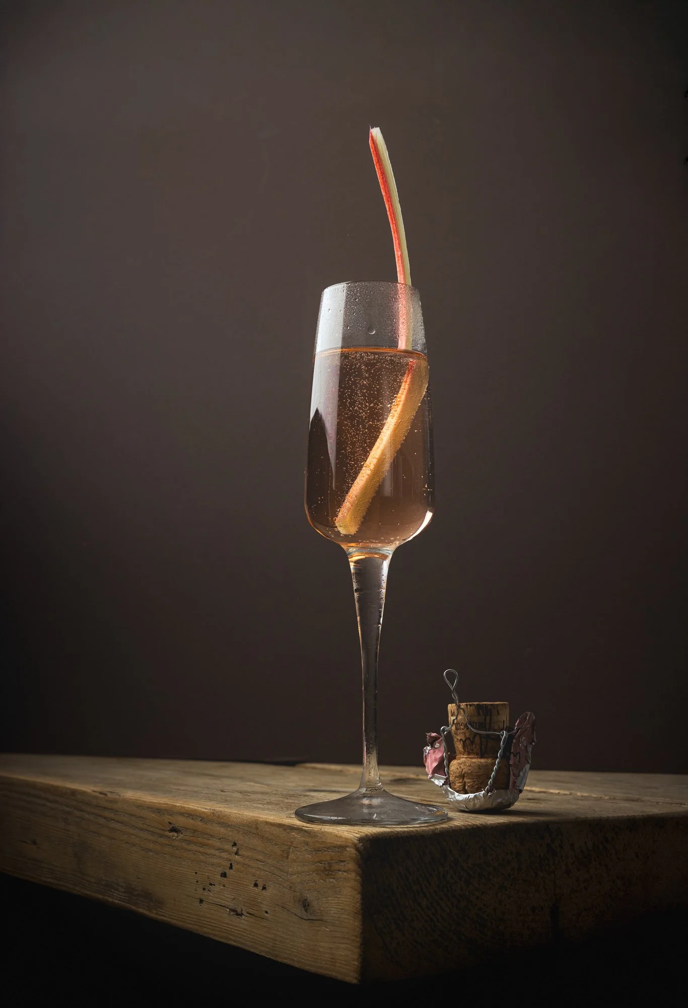 Commercial cocktail photograph of a pale sparkling drink in a tall stemmed glass with a long curved citrus or rhubarb garnish, presented on a rustic wooden surface against a softly graduated background.