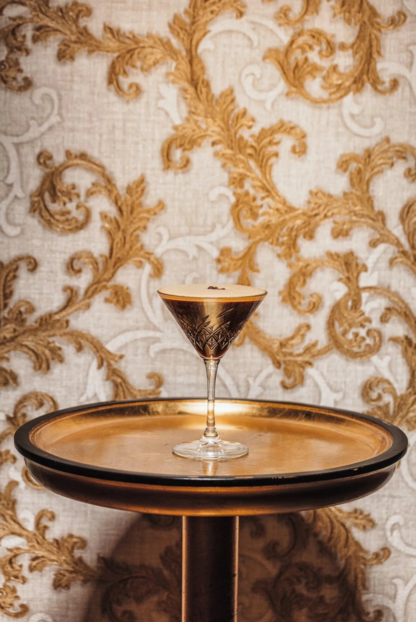Commercial cocktail photograph of a pale martini-style drink presented in a stemmed glass on a circular serving tray, using warm tones and patterned wallpaper to give the image a refined hospitality setting