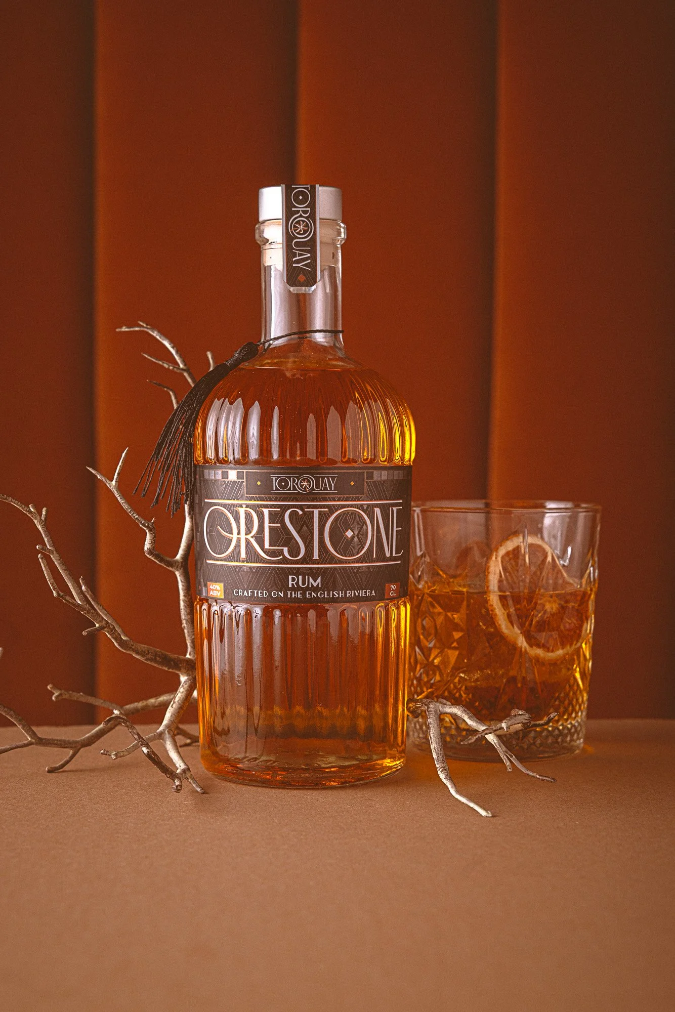 Commercial product photograph of an Orestone rum bottle with a filled tumbler and sculptural branches, using a warm terracotta set and controlled reflections to present the amber spirit, bottle shape and label clearly.