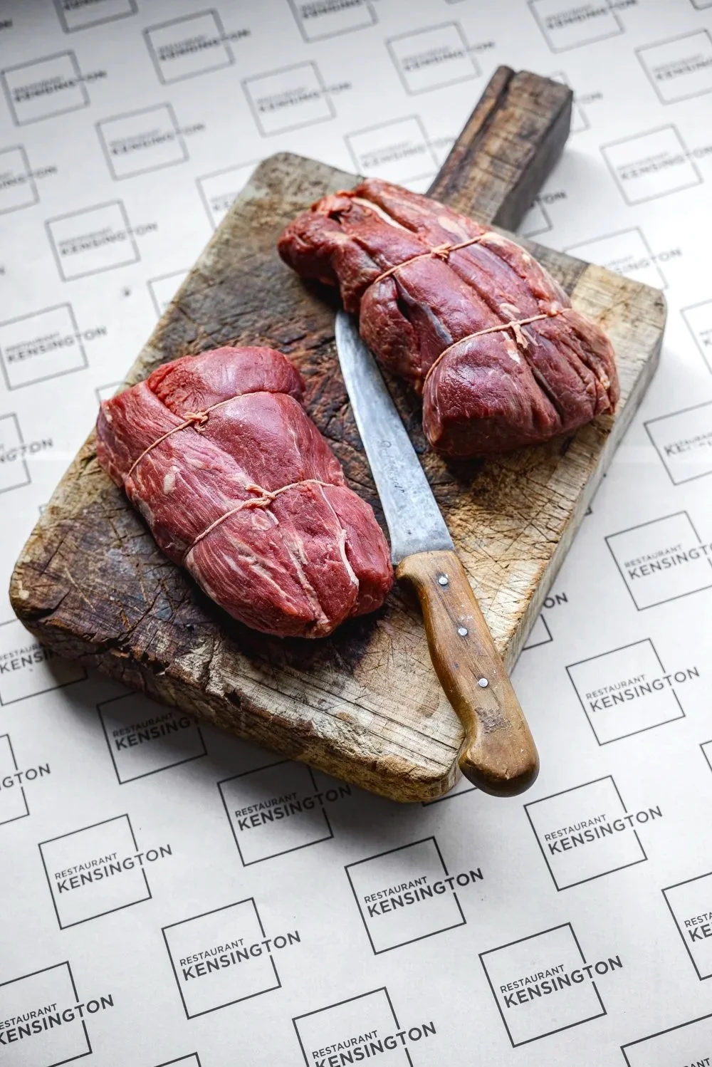 Commercial product photograph of two raw steaks presented with a knife on branded butcher paper, combining clear product detail with a practical preparation surface for Kensington restaurant butcher on line  in Devon.