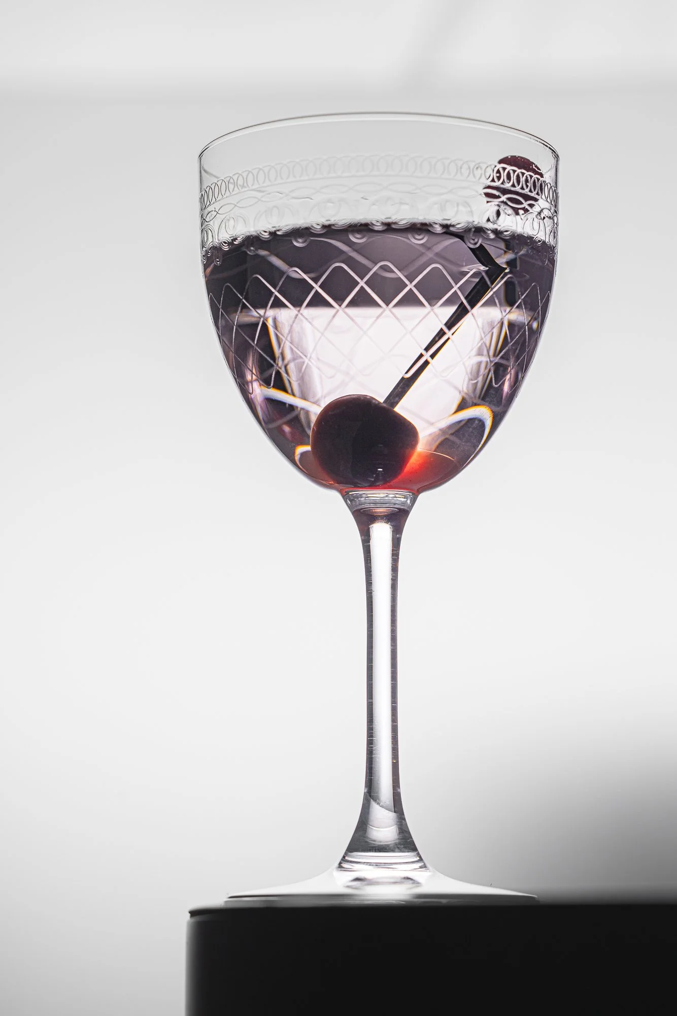Studio cocktail photograph of a clear cocktail with a dark cherry in an etched stemmed glass, isolated against a pale background with precise lighting to reveal the glass pattern, liquid clarity and garnish.