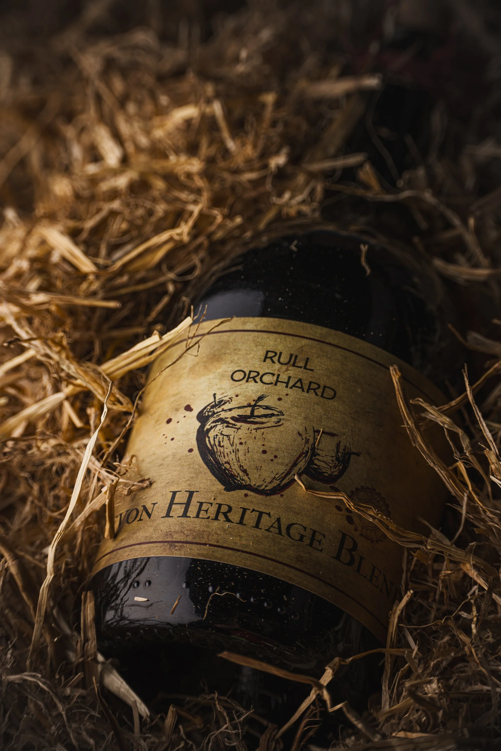Close commercial product photograph of a Rull Orchard heritage cider bottle resting among straw, using warm directional light to emphasise the illustrated label, glass surface and connection with rural Somerset production.