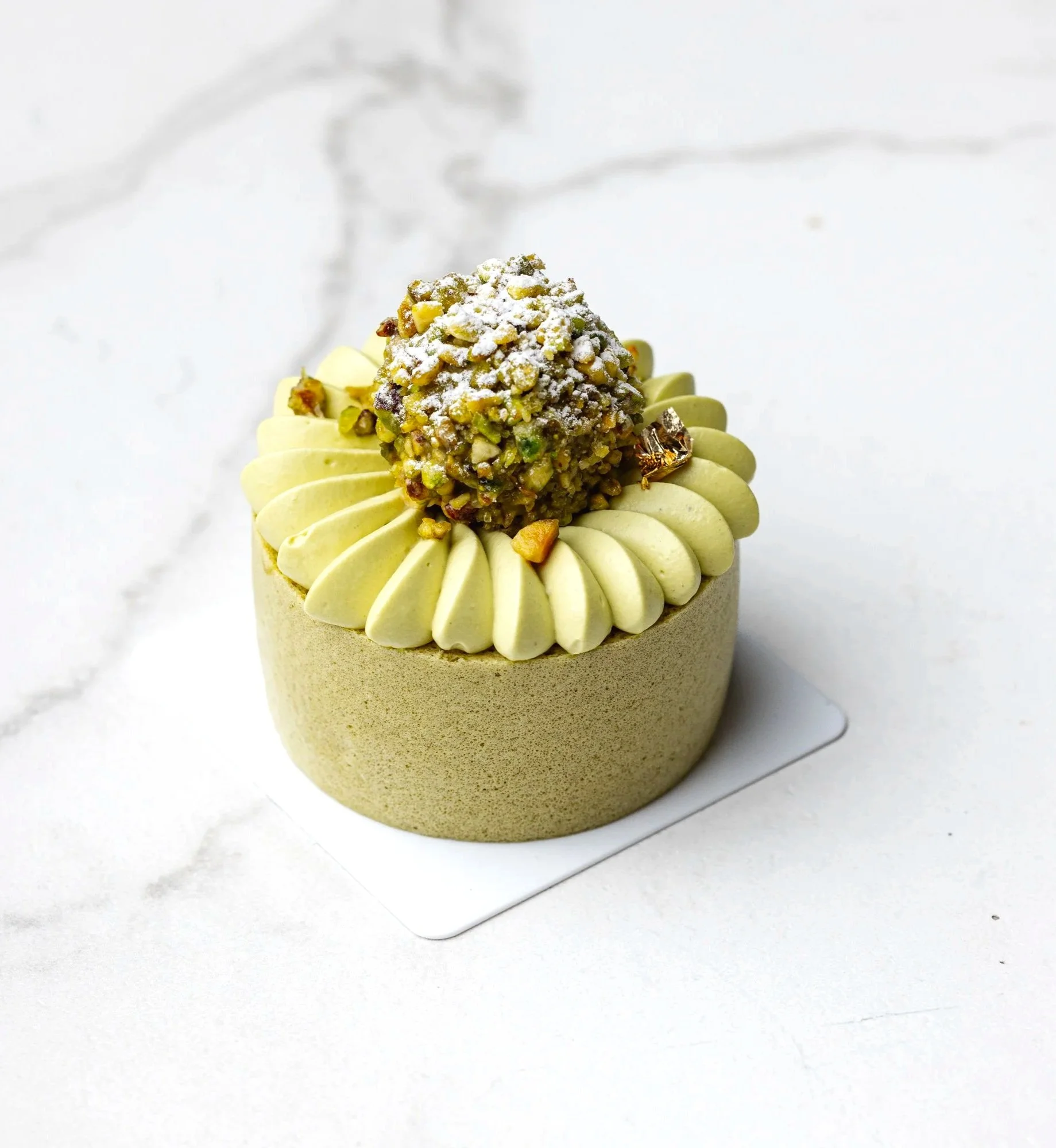 Minimal commercial pastry photograph of a small green individual cake topped with piped cream and pistachio pieces, presented on a pale marble surface to highlight its precise finish and layered construction.
