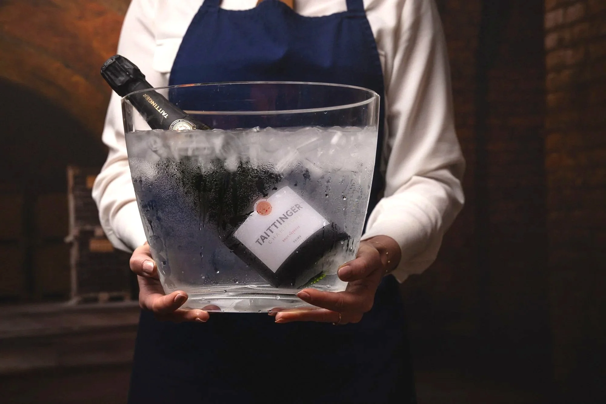 Hospitality photograph of a server holding a clear ice bucket containing a chilled Taittinger Champagne bottle, using condensation, ice and formal uniform details to communicate premium drinks service for Posh Nosh Devon.