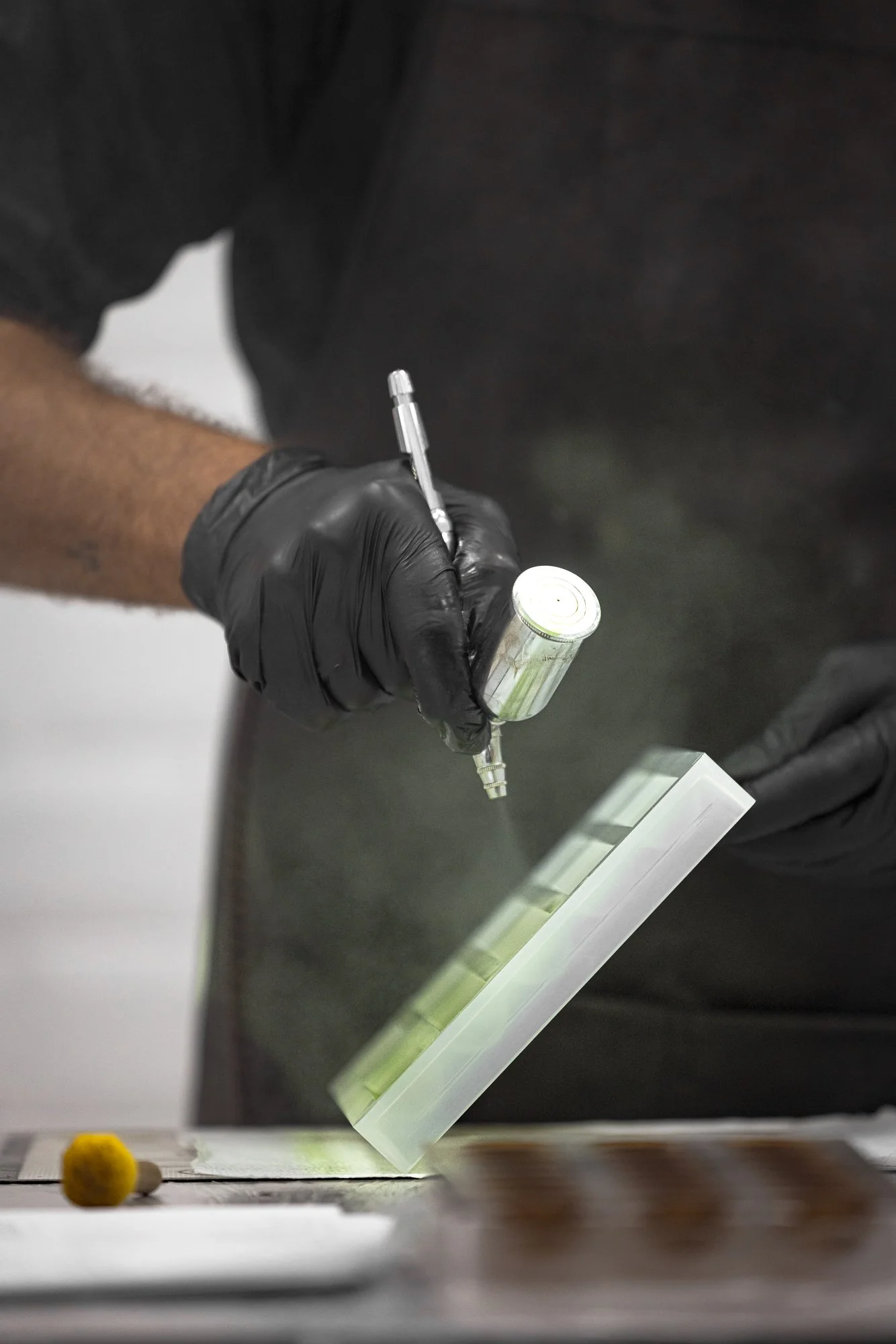 Chocolate production photograph showing a gloved pastry chef spray-coating a mould with coloured cocoa butter, capturing the specialist technique, mist and controlled working environment for client or chocolatier.