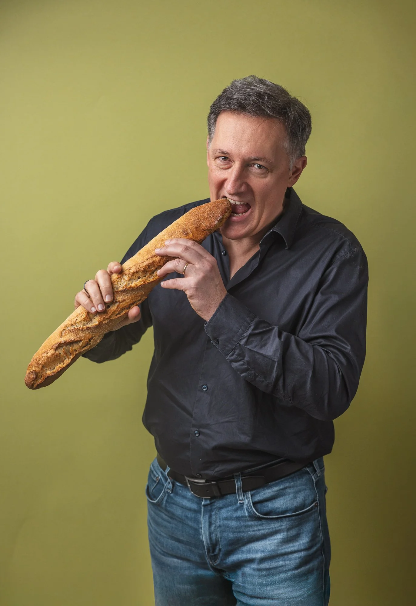 Playful editorial portrait of a food-industry professional biting into a long baguette against a muted green background, using humour and a simple studio composition to show personality alongside his connection with bread and food.