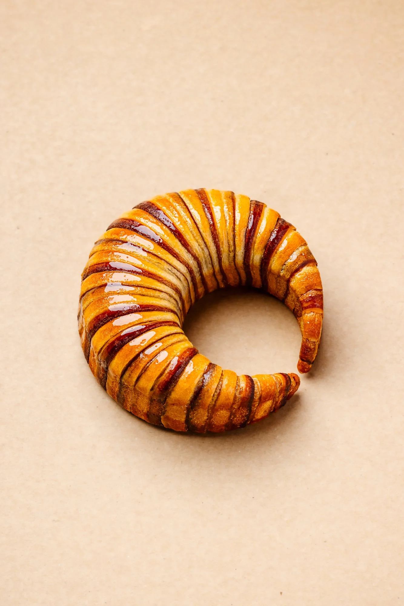Minimal commercial photograph of a glazed croissant by Antonio Bachour  standing upright against a warm cream background, using controlled light to define the layered surface and circular form