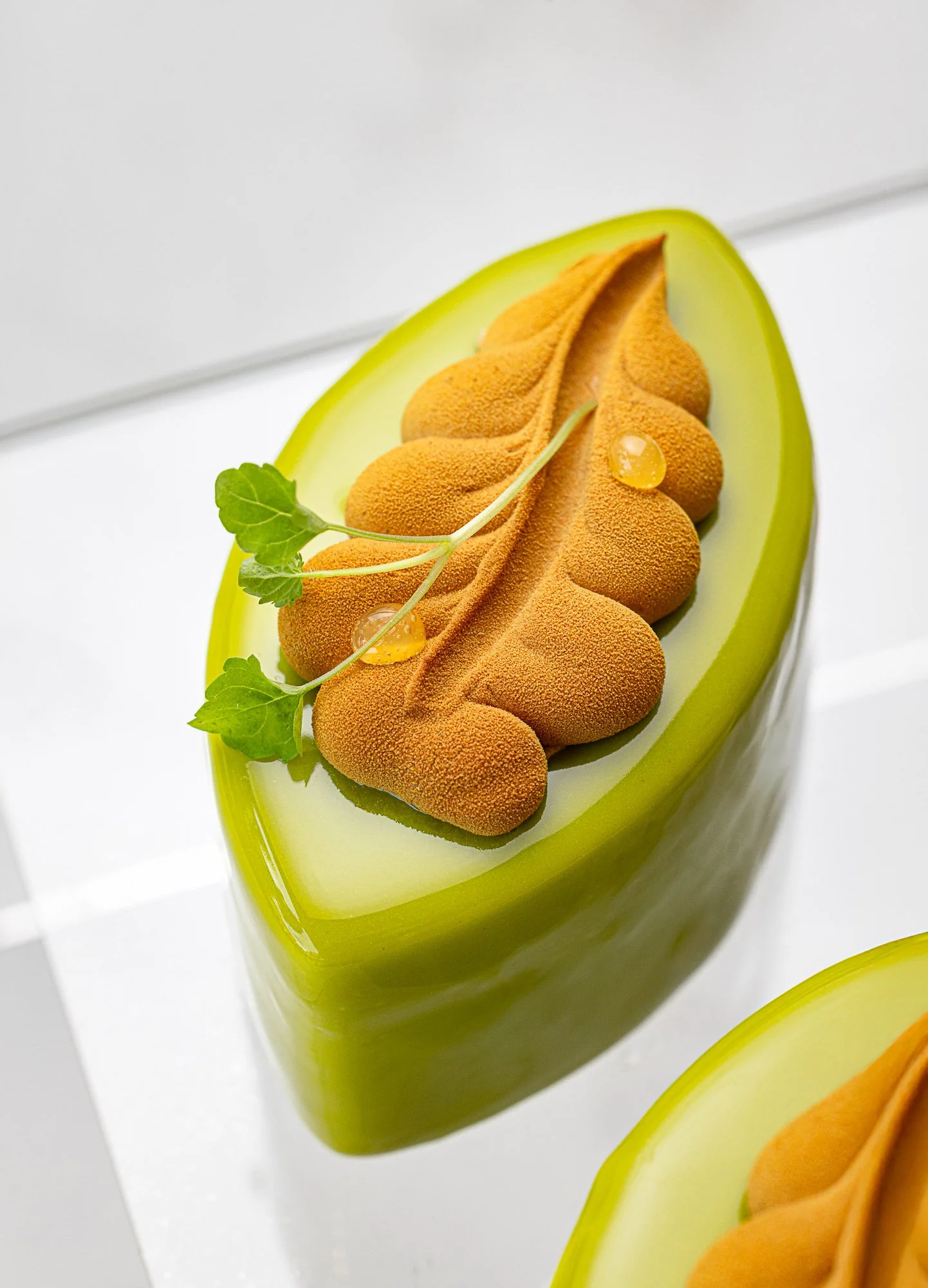 Commercial pâtisserie photograph of a glossy green oval dessert topped with biscuit-like elements and herbs, presented on a white background to emphasise its sculptural shape, layers and refined finish.