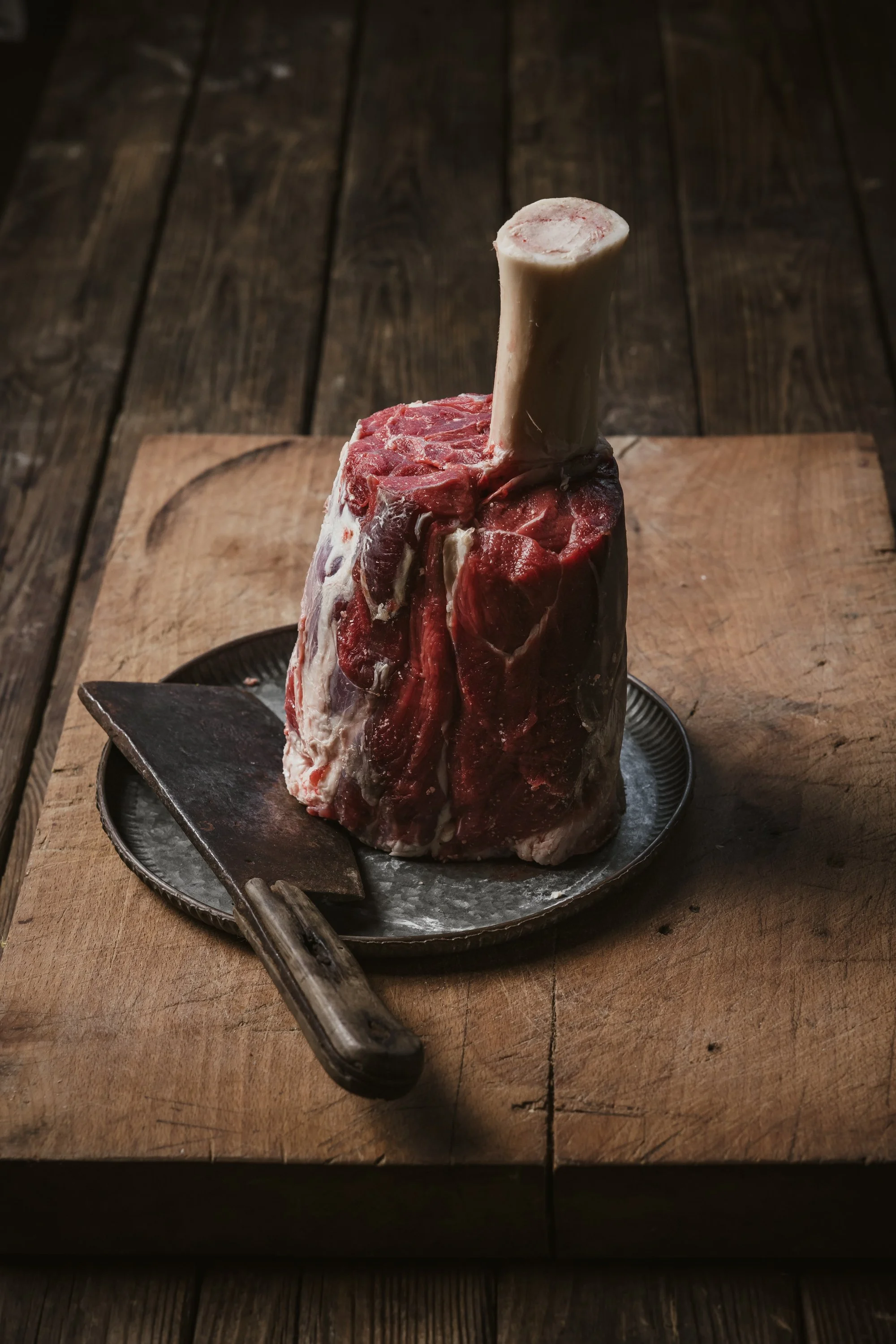 Commercial food photograph of a thick bone-in shin cut standing upright on a dark board with a knife, using rustic styling and side lighting to emphasise the meat’s structure and texture for Wonky Horn shop online in Somerset.