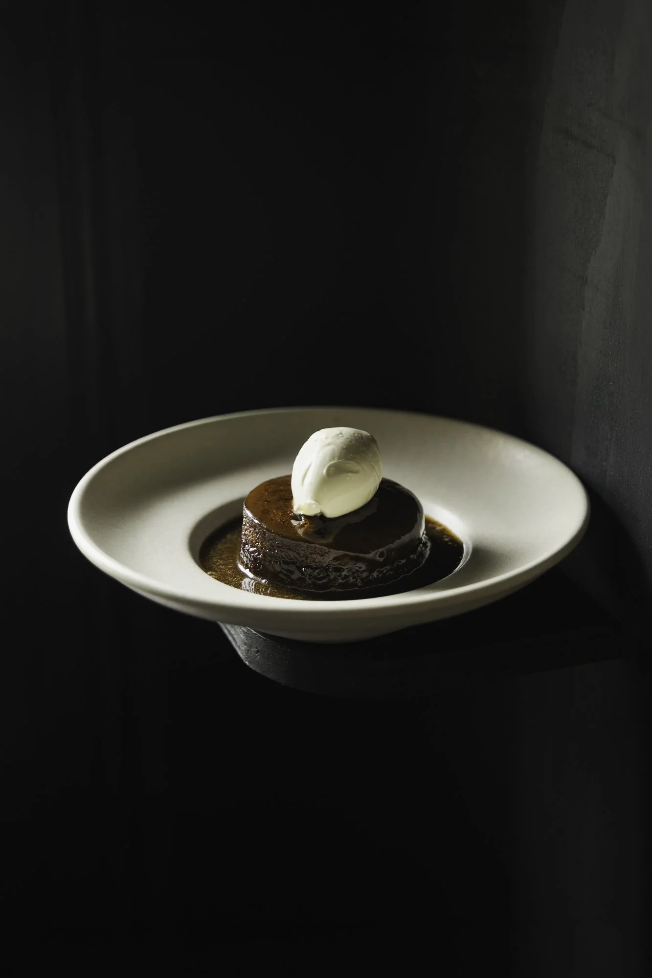 Commercial food photograph of sticky toffee pudding presented in a wide white bowl against a dark background, with focused lighting emphasising the shape, sauce and contrasting scoop for The George public house website in Somerset