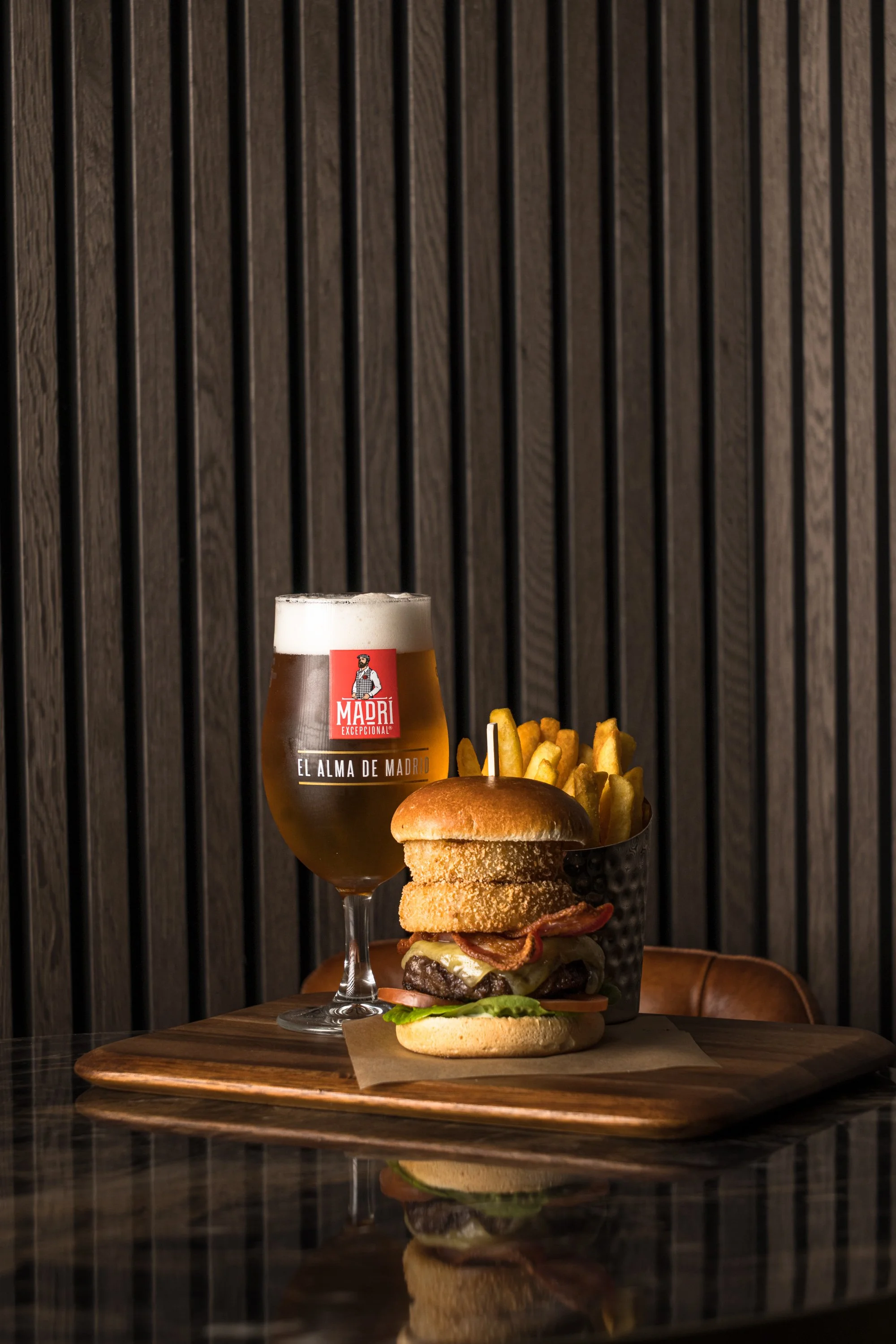 Commercial restaurant photograph of a stacked burger served with chips and a glass of beer against dark vertical panelling, using controlled directional lighting for Exeter Golf club in Devon