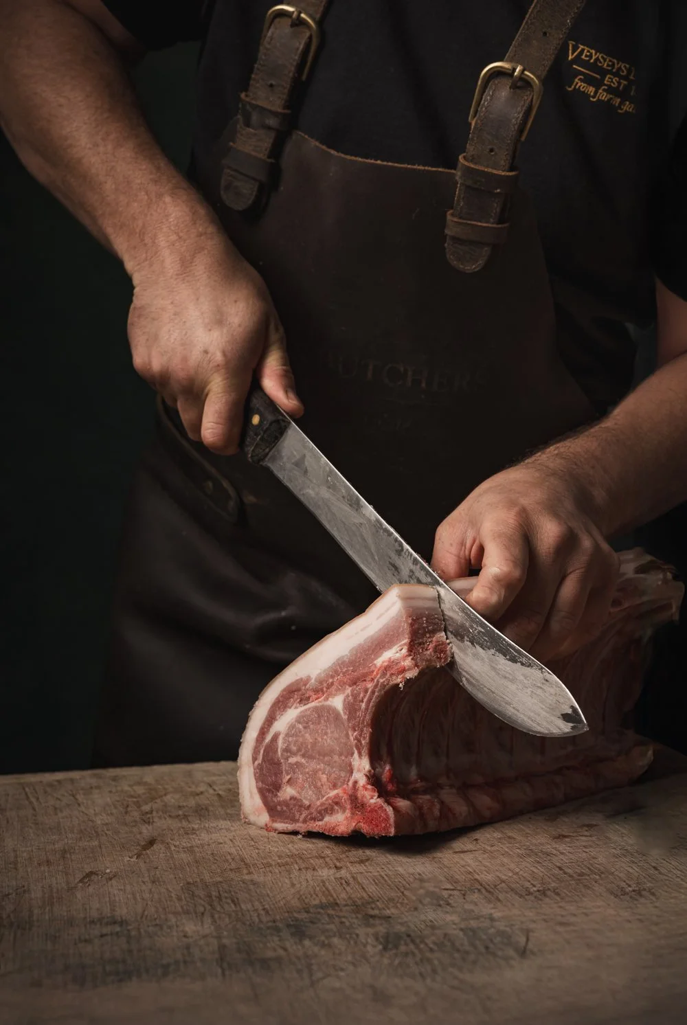 Commercial butchery photograph showing a butcher from Veyseys butcher  cutting through a thick pork chop with a large knife, using dark directional lighting to emphasise the hands, raw meat and practical working environment.