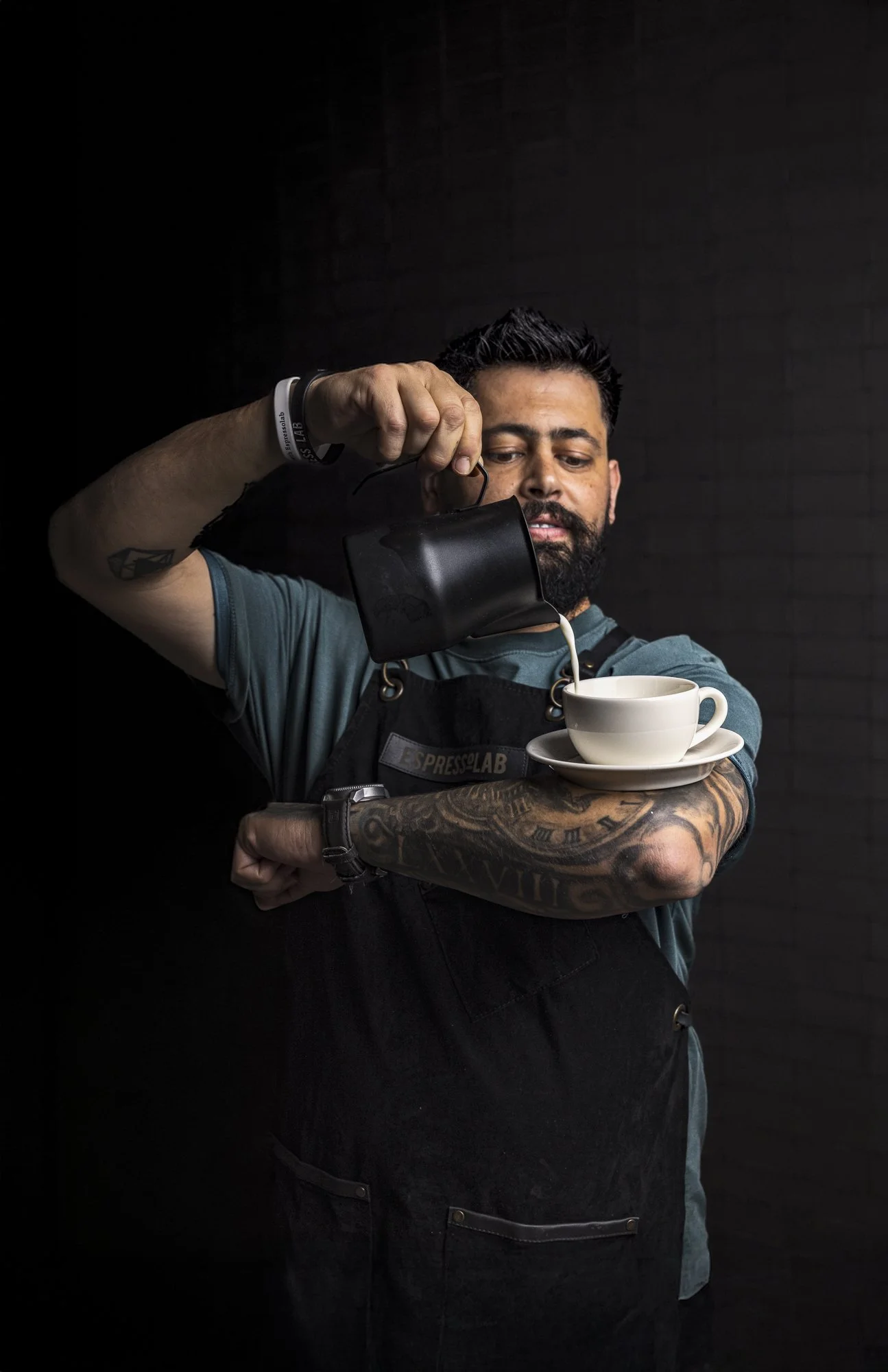 Commercial barista portrait showing a tattooed coffee professional pouring steamed milk or coffee from a black jug into a white cup, using a dark background to focus attention on the person, gesture and drink service.