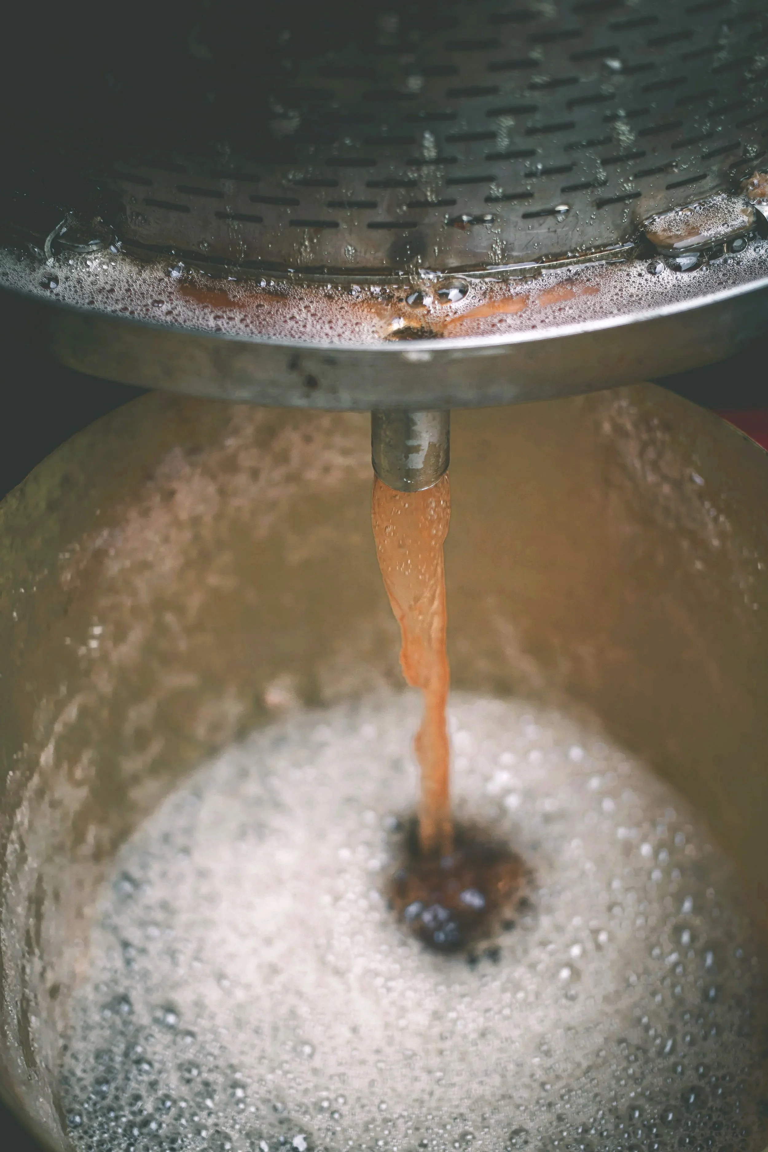 Close production photograph of freshly pressed amber liquid flowing from stainless-steel equipment into a foaming collection vessel, documenting the extraction process used to produce cider in Somerset.