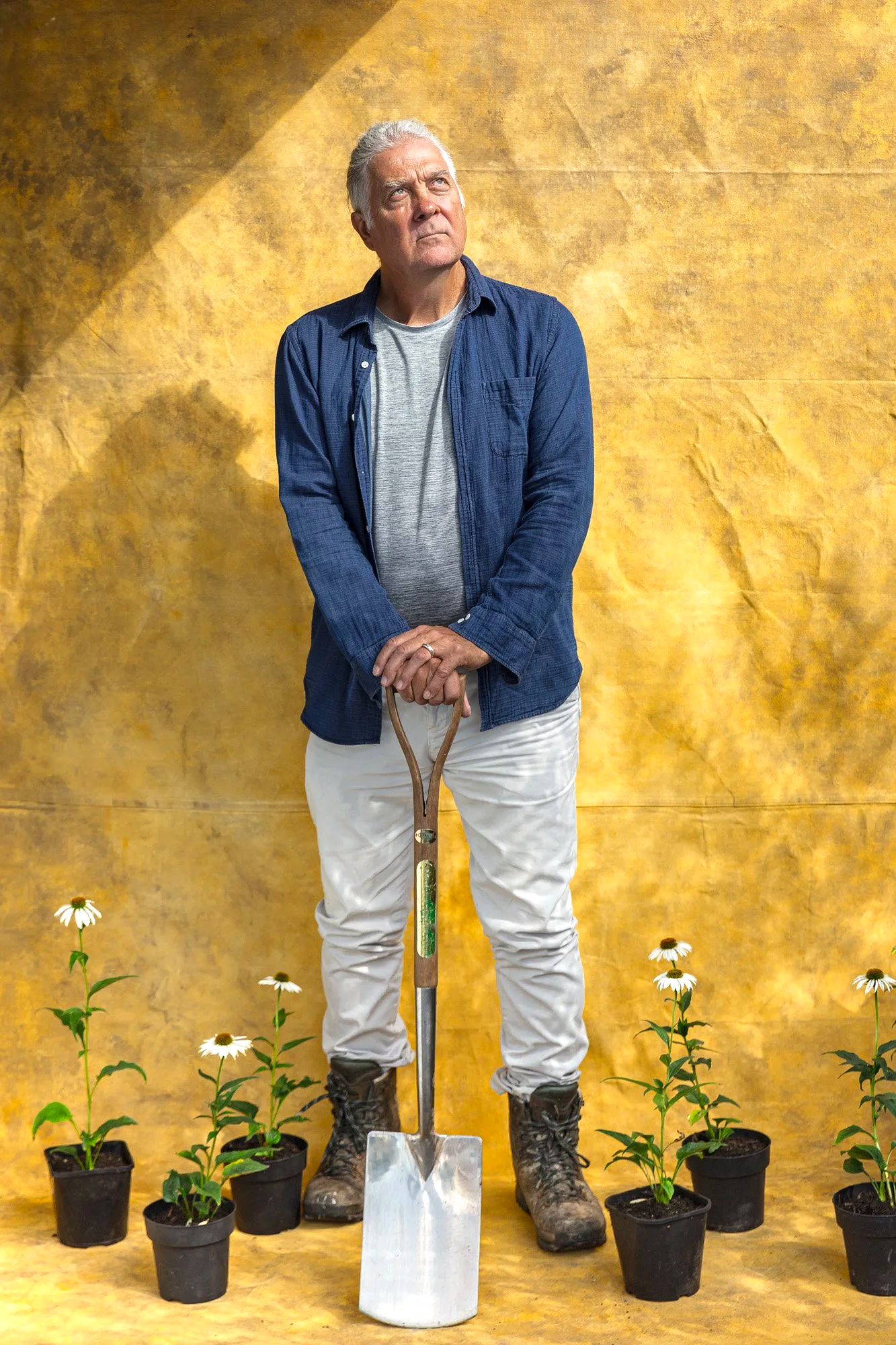 Character-led editorial portrait of an older man standing with a garden spade between small plants against a textured gold wall, using an understated pose to connect the subject with horticulture, growing or land-based work.