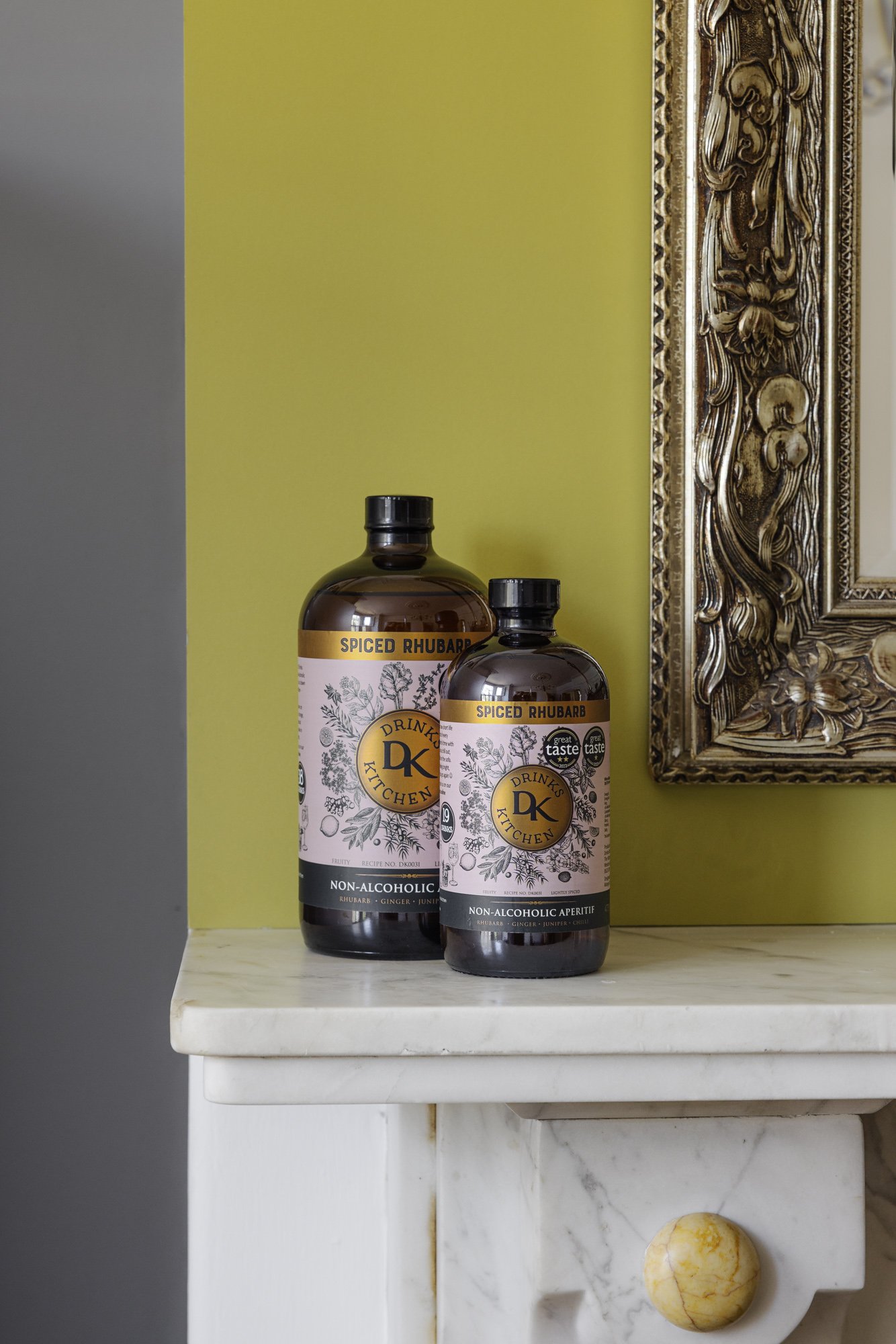 Commercial product photograph of two Drink Kitchen bottles displayed on a white mantel against a yellow-green wall, combining clear packaging detail with an understated domestic setting for Drink Kitchen in Bristol