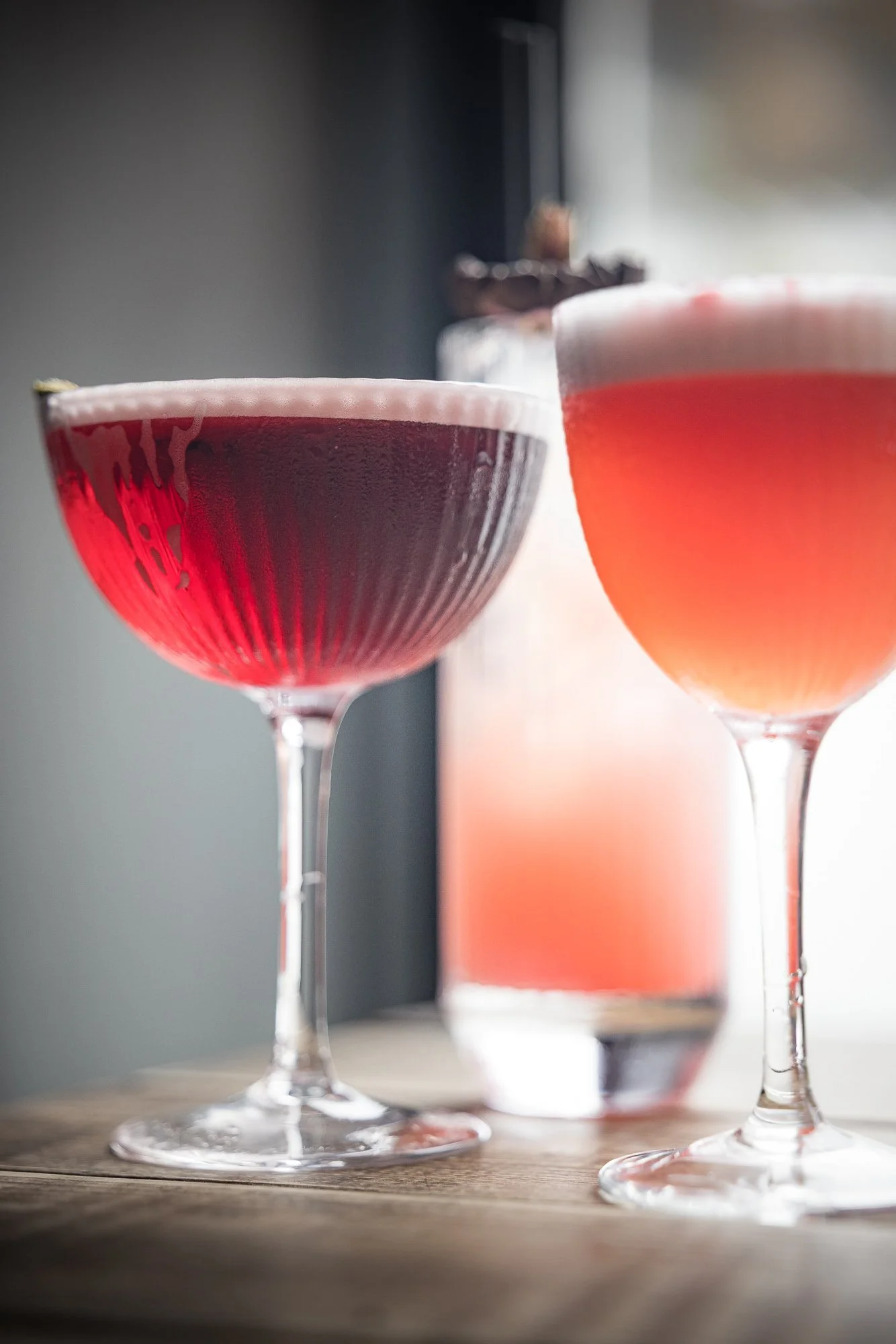 Commercial drinks photograph of two vivid red cocktails in stemmed glasses beside a softly focused matching bottle, using bright window light to reveal the translucent colour, glass reflections and contrasting silhouettes.