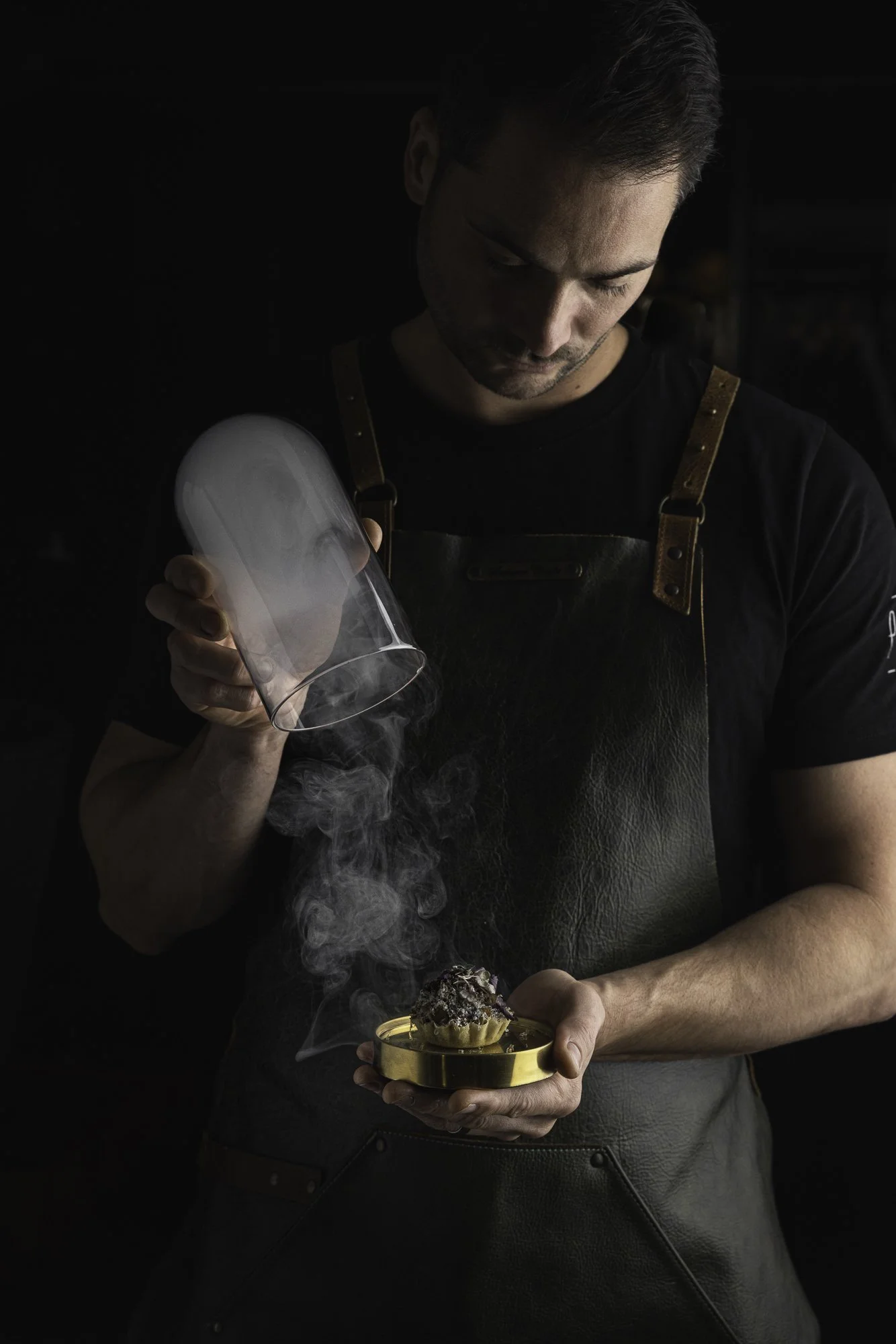Restaurant food photography showing a chef lifting a smoke-filled cover from tartlet held at chest height, capturing the theatrical presentation and working hands for Pasture in Cardiff.
