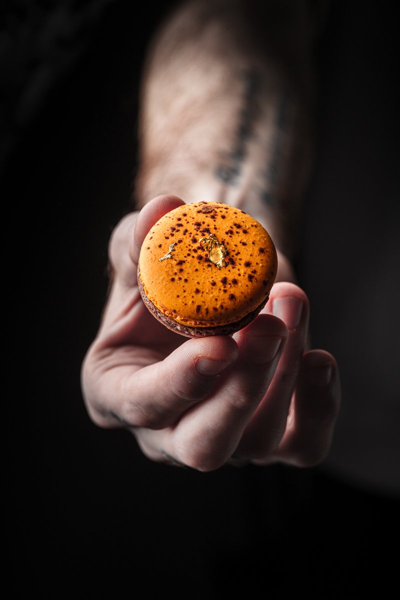 Close commercial pastry photograph of a vivid orange macaron held carefully in one hand against a dark background, using directional light to emphasise the shell, filling and decorative speckled finish.
