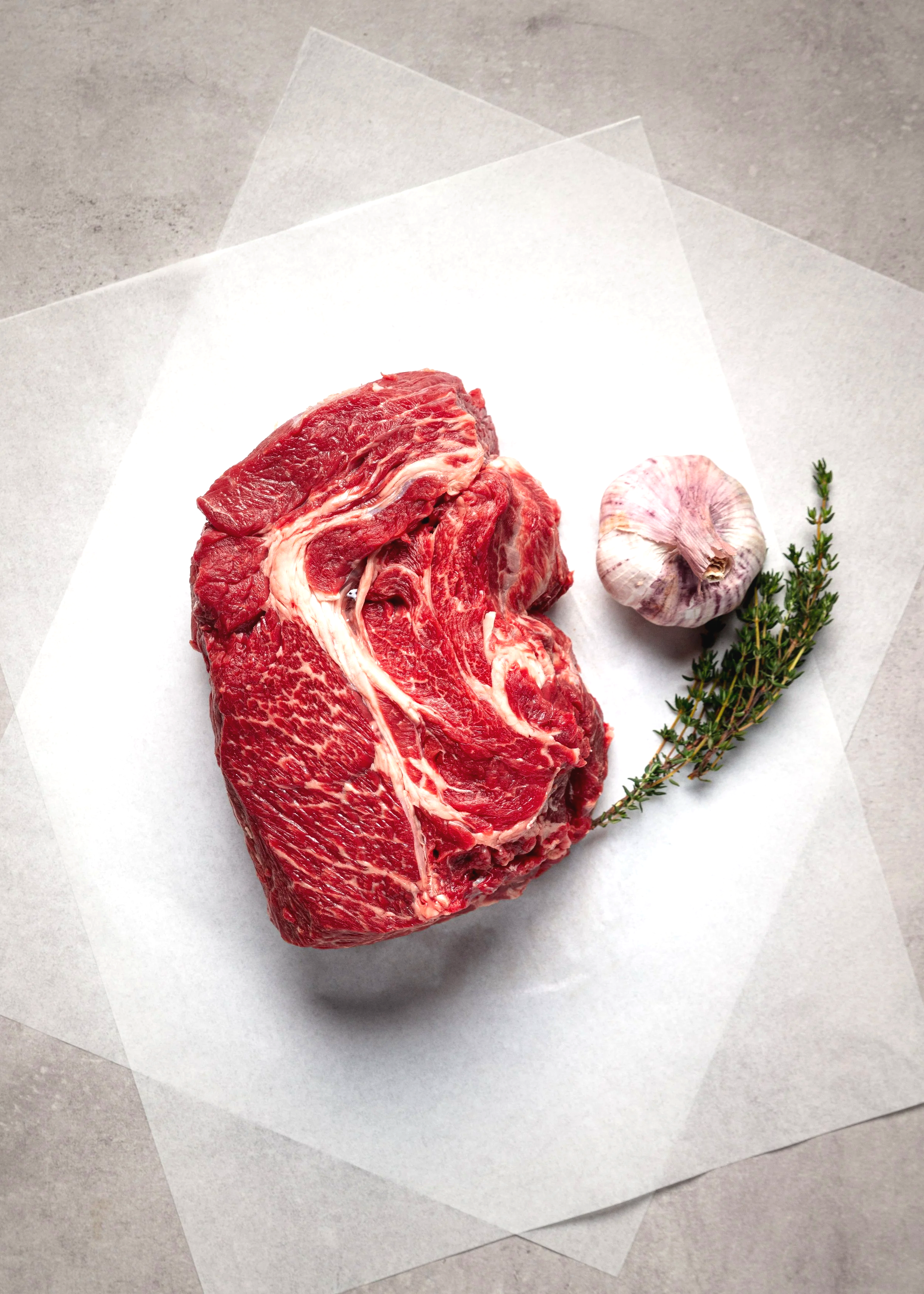 Top-down commercial meat photograph of a raw ribeye steak on white butcher paper with garlic and rosemary, using clean lighting to show its marbling, colour and fresh-cut texture.