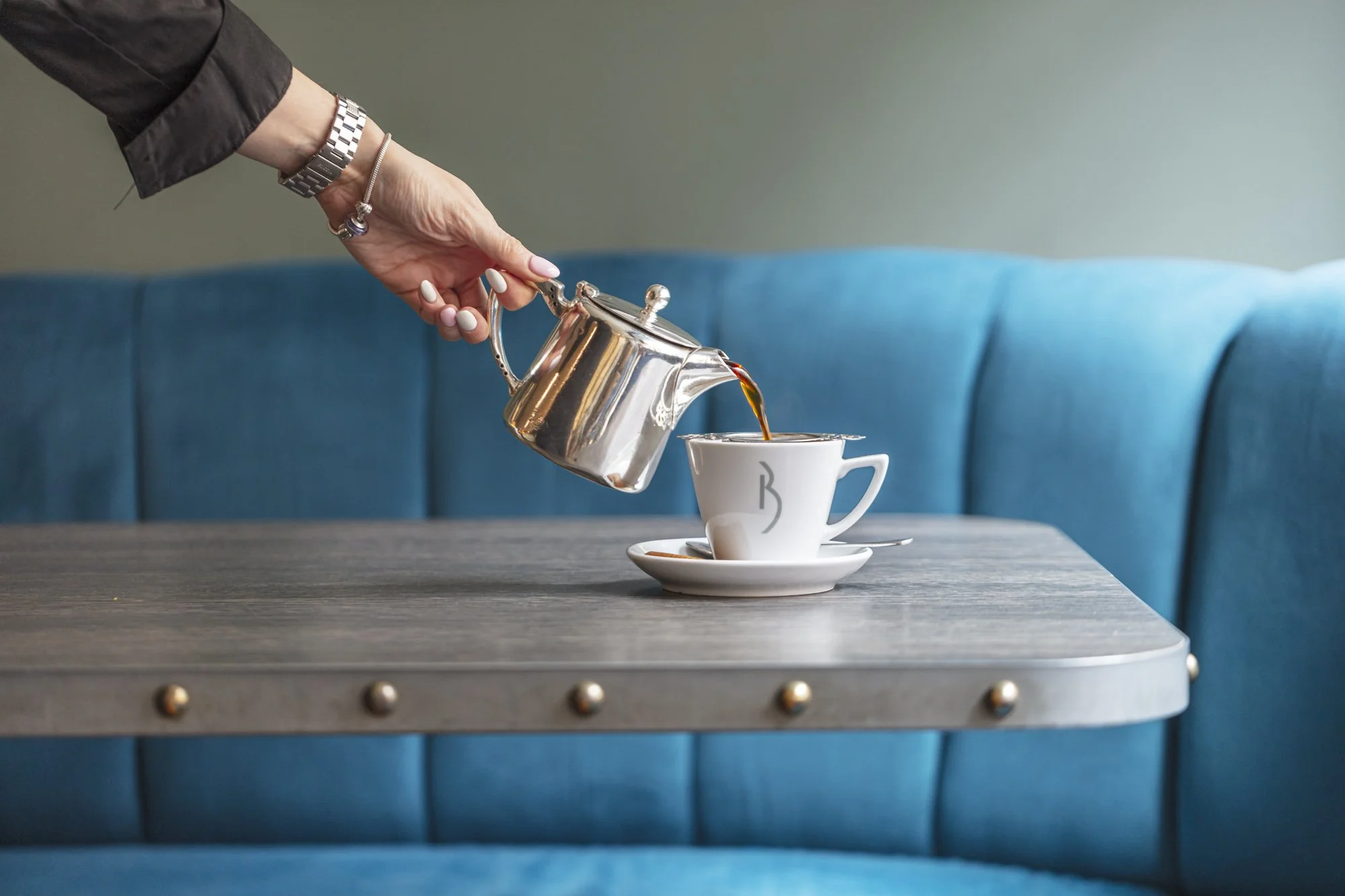 Hospitality photograph of tea being poured from a polished metal pot into a white cup at a café table, using a bright blue banquette and simple setting to communicate attentive table service.