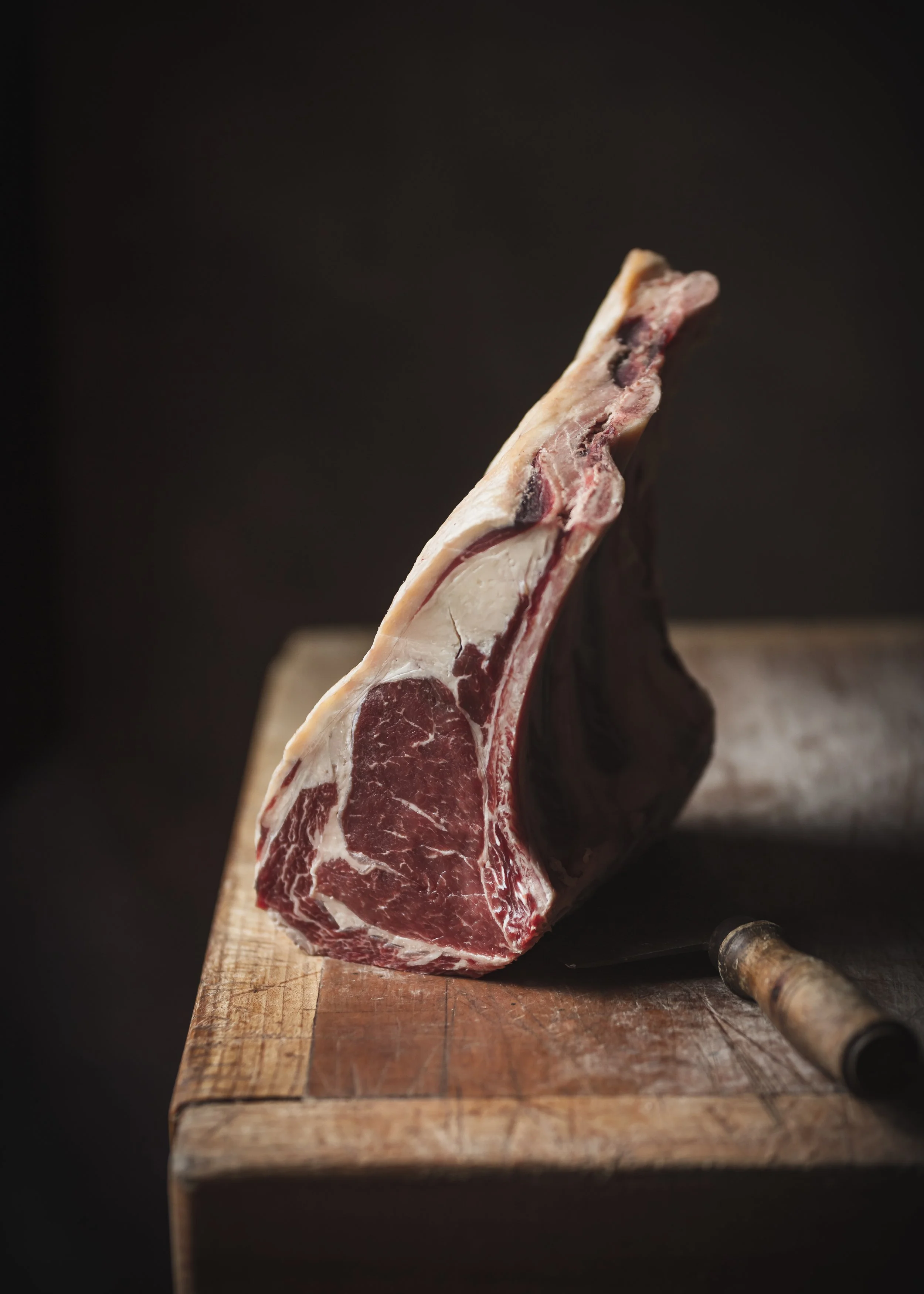 Commercial meat photograph of a single bone-in cut on a wooden block against a dark neutral background, using simple styling and directional light to emphasise its shape, marbling and natural texture.