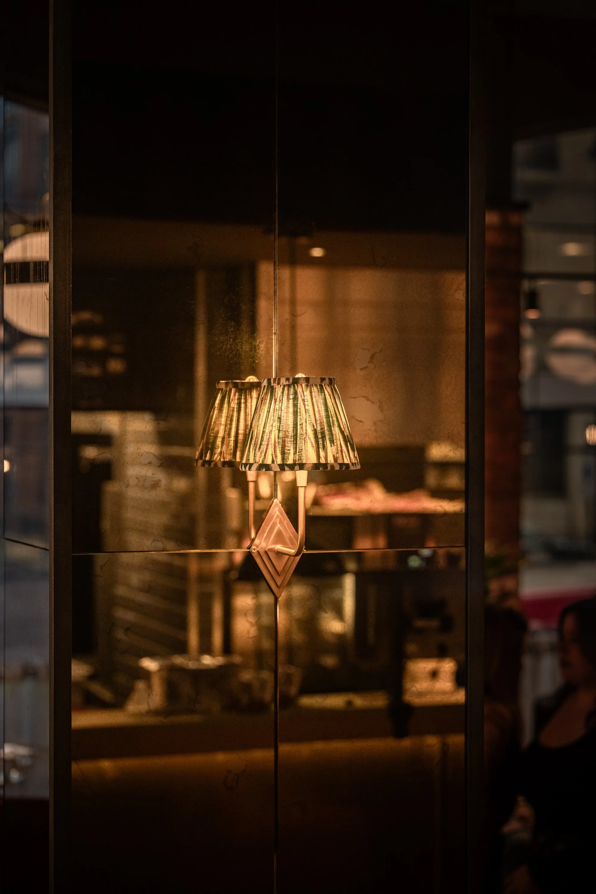 Atmospheric interior detail for Pasture Restaurant Group, showing a patterned table lamp reflected in dark glass with warm lighting and furnishings softly visible behind it.