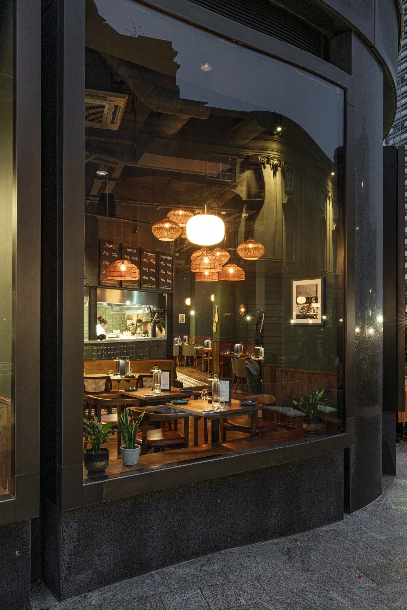 Evening hospitality photograph through the window of The Cow & Sow in Birmingham, showing dining tables, warm pendant lights, reflections and the open kitchen beyond.