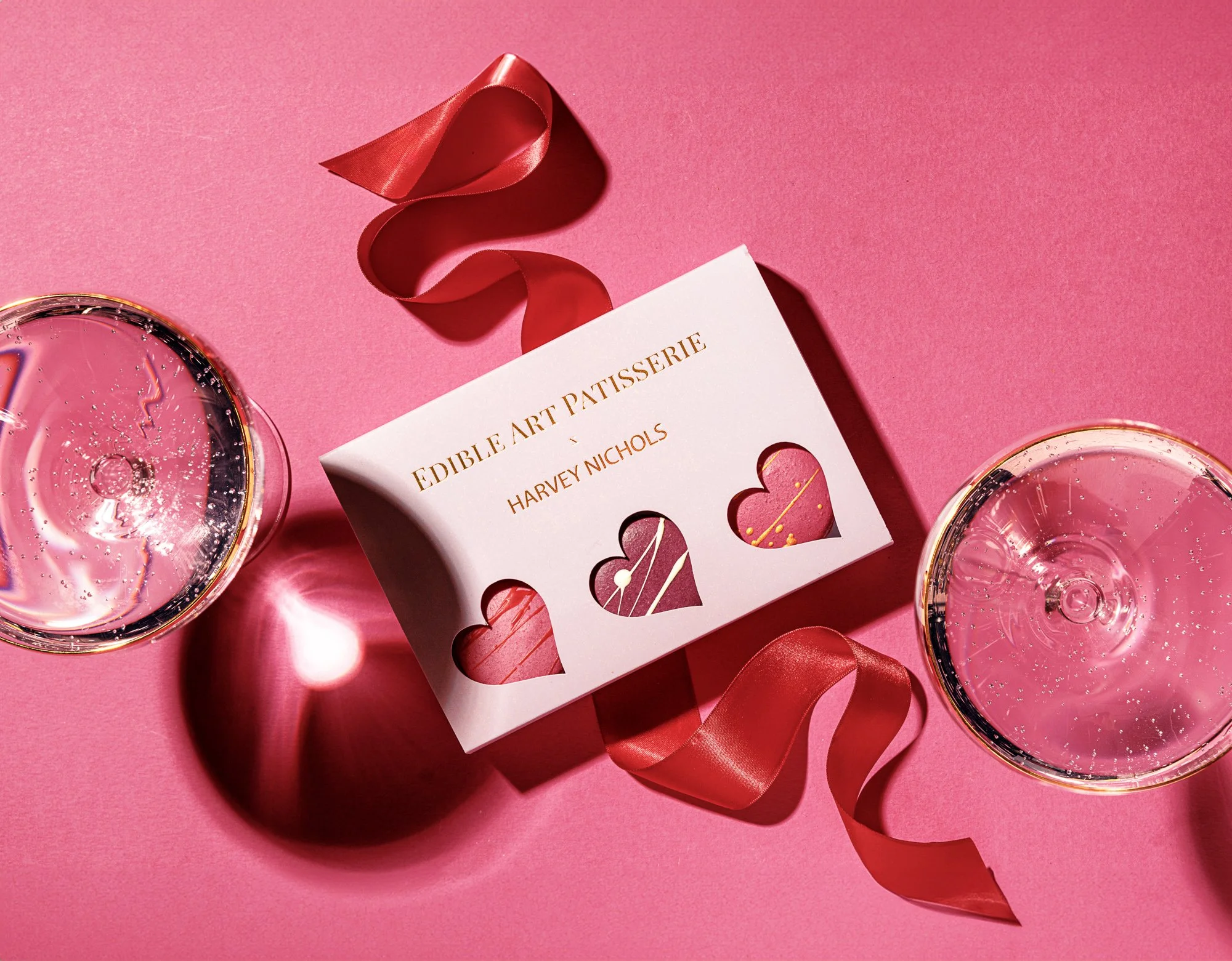 Commercial Valentine’s product photograph of a pink heart-themed chocolate gift box surrounded by ribbon and reflective glasses, using strong colour blocking and overhead composition by Damien Wager for Harvey Nichols retail.