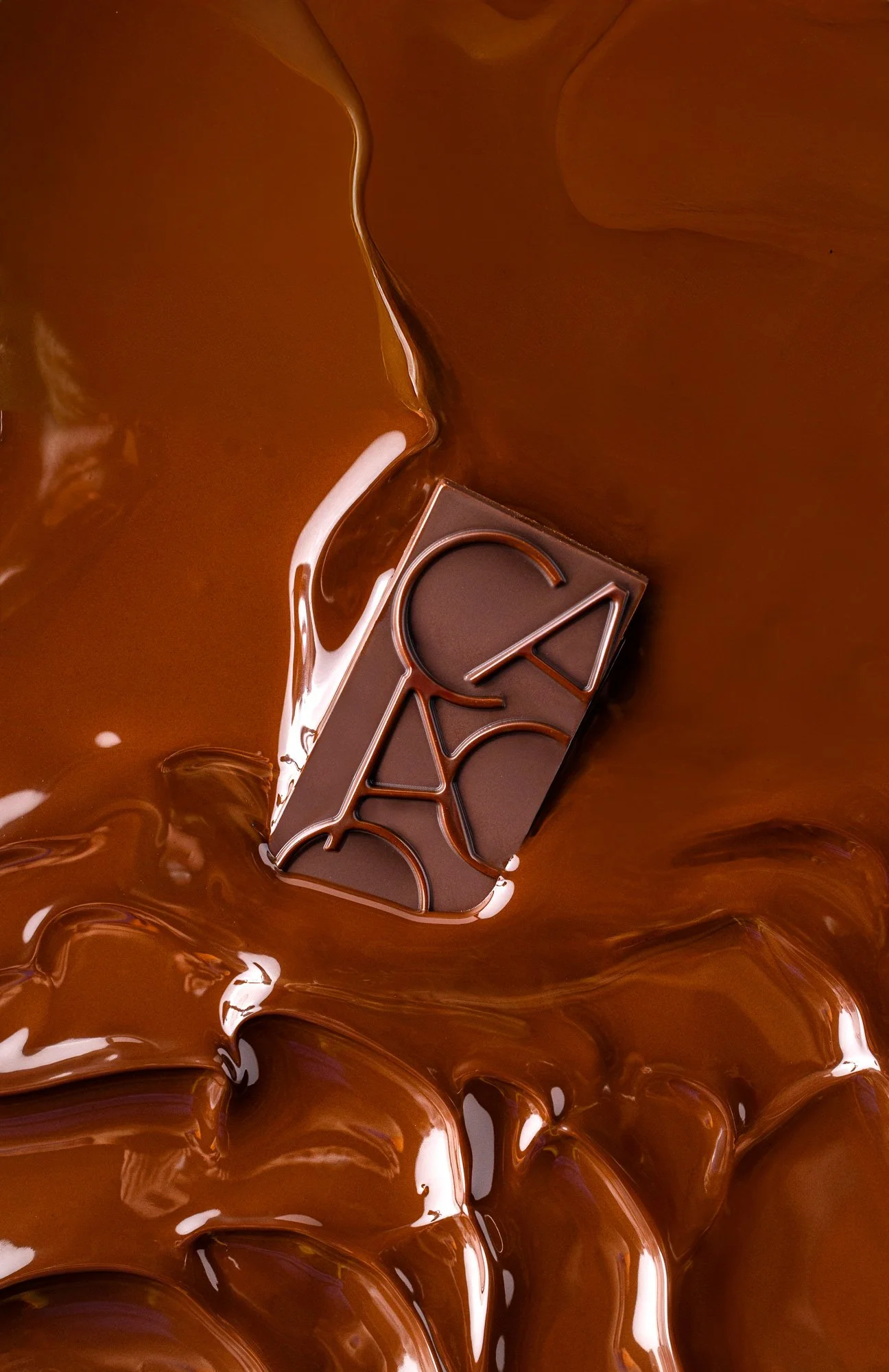 Commercial chocolate photograph of a geometric chocolate bar partially submerged in glossy melted chocolate, using a close composition and warm brown palette to emphasise fluid texture and moulded detail.