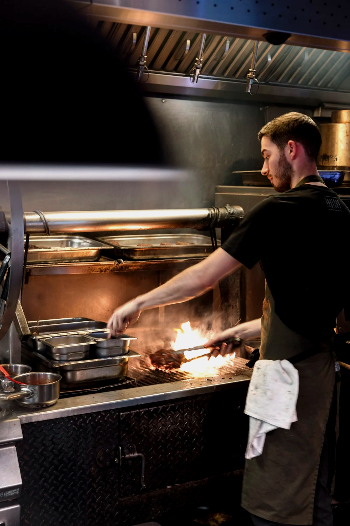 Documentary kitchen photograph of a chef working over a burst of flame on the restaurant stove, capturing the heat, concentration and pace of professional service for Pasture in Cardiff.