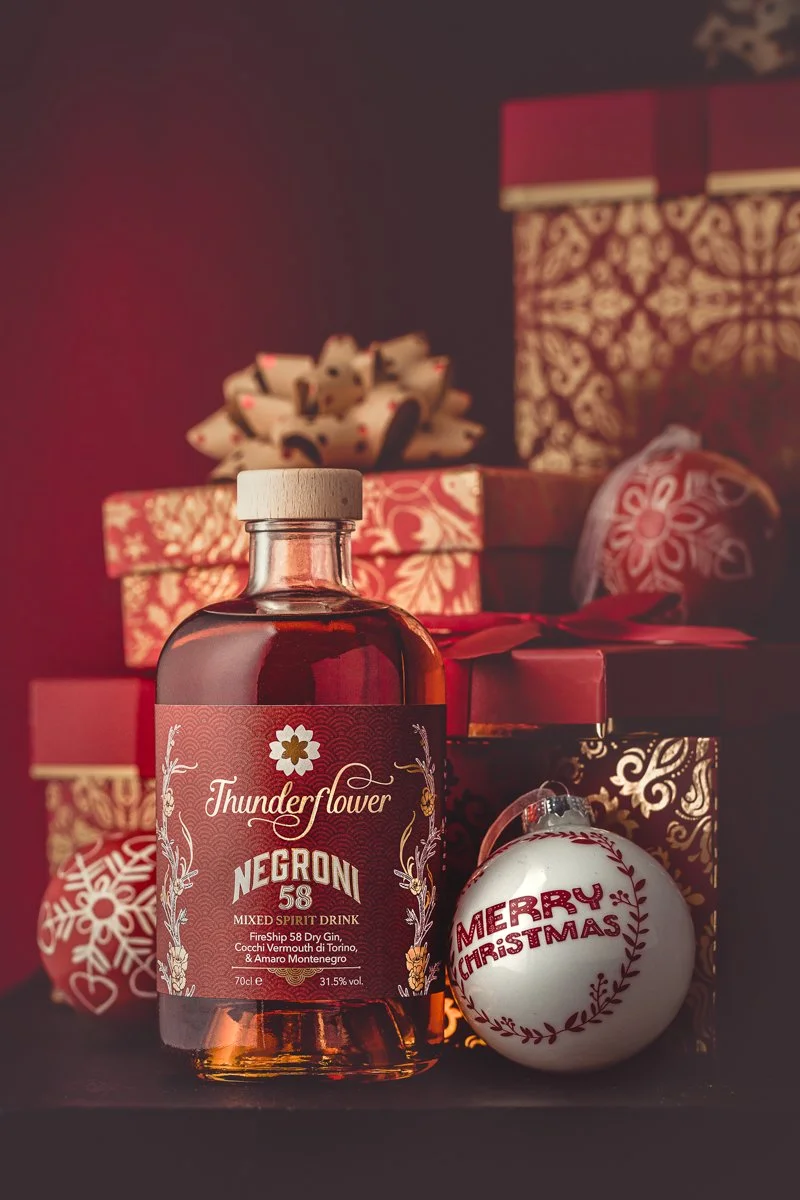 Seasonal commercial photograph of a Thunderflower spirit bottle arranged with wrapped Christmas gifts and a festive decoration, using a rich red set to present the product as a premium seasonal gift.