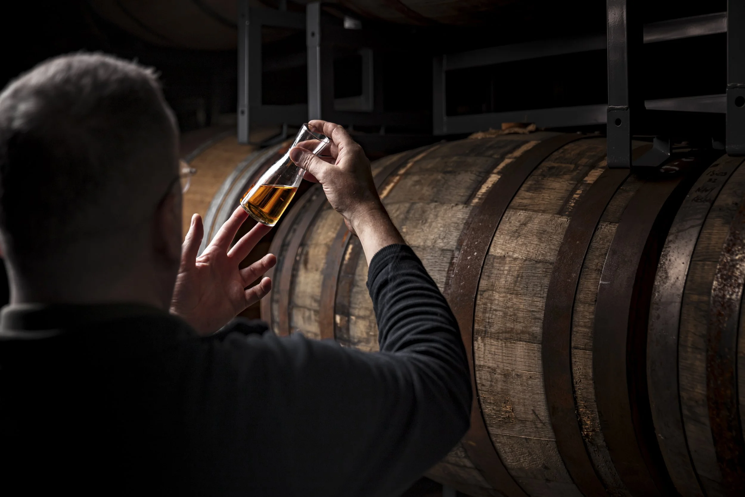 Documentary distillery photograph showing a producer holding a glass of amber spirit beside a wooden barrel, capturing the sampling and maturation process in a dark working cellar for distillery in Exmoor.