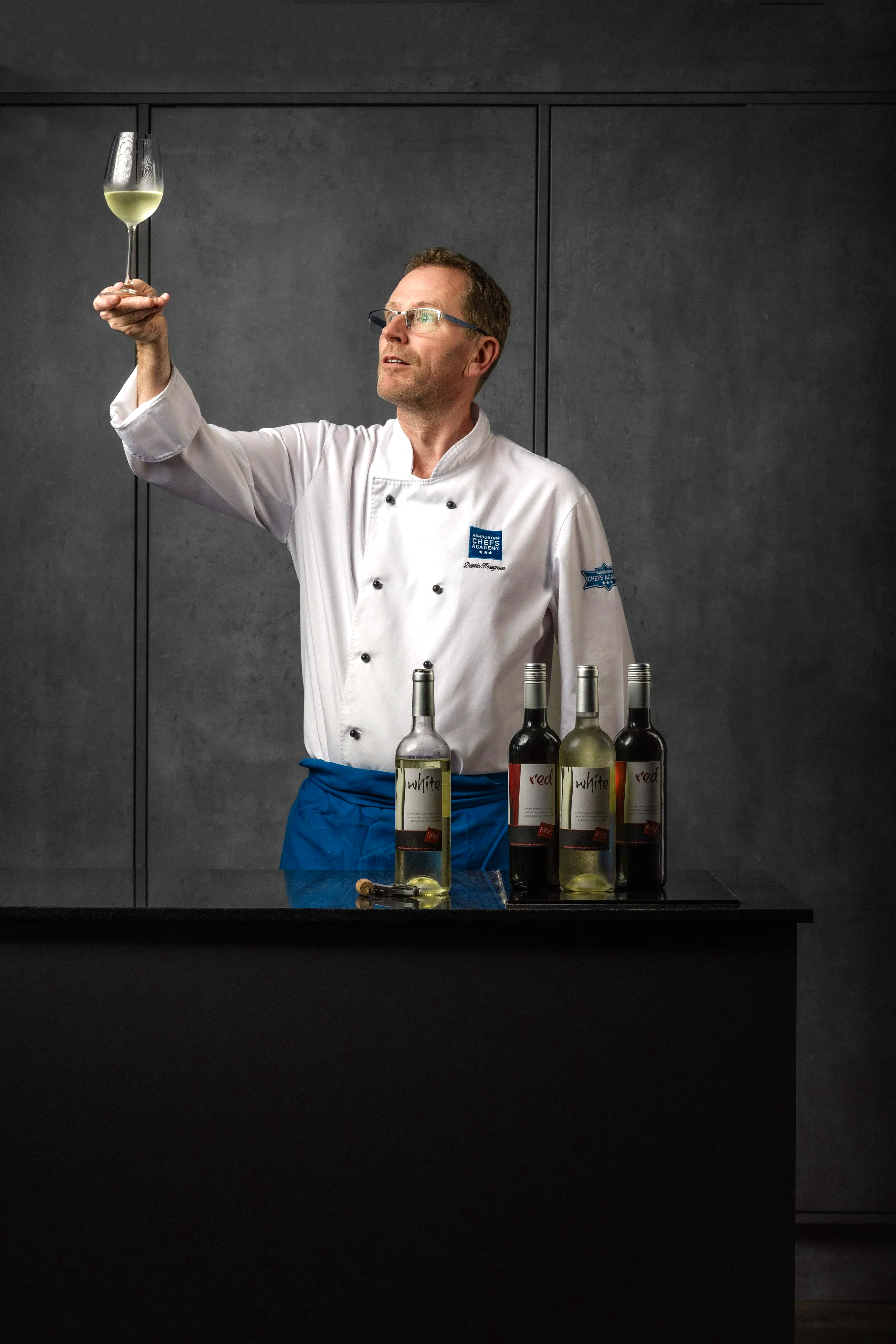 Editorial portrait of a wine professional raising a glass towards the light above a row of bottles, connecting the person with tasting, assessment and the drinks industry for Ashburton Cookery School communication.
