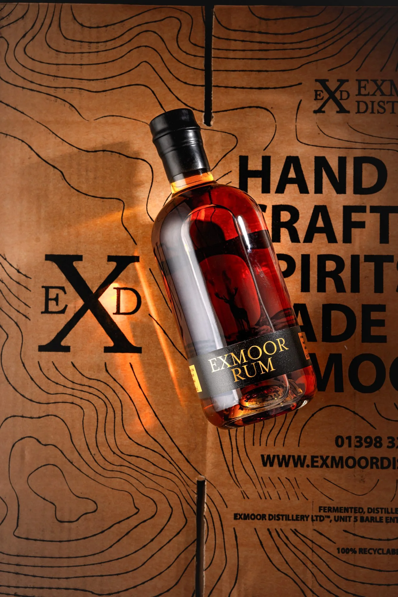 Commercial promotional photograph of an Exmoor Distillery rum bottle arranged diagonally over branded map graphics and typography, combining product photography with a bold campaign layout linked to the spirit’s regional identity.
