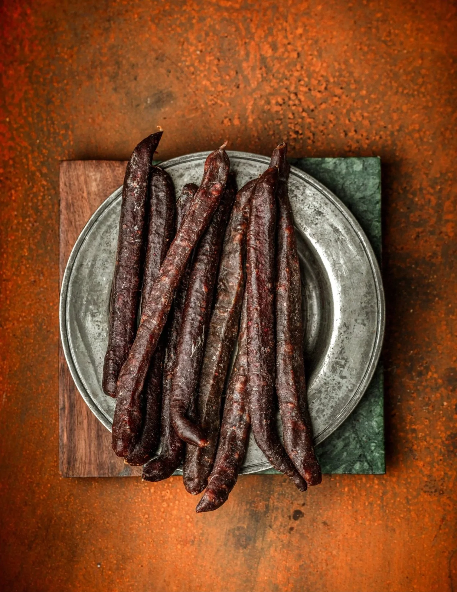 Top-down commercial food photograph of dark artisan sausages arranged on a metal plate over a rust-coloured surface, using restrained props and strong texture for a butcher .