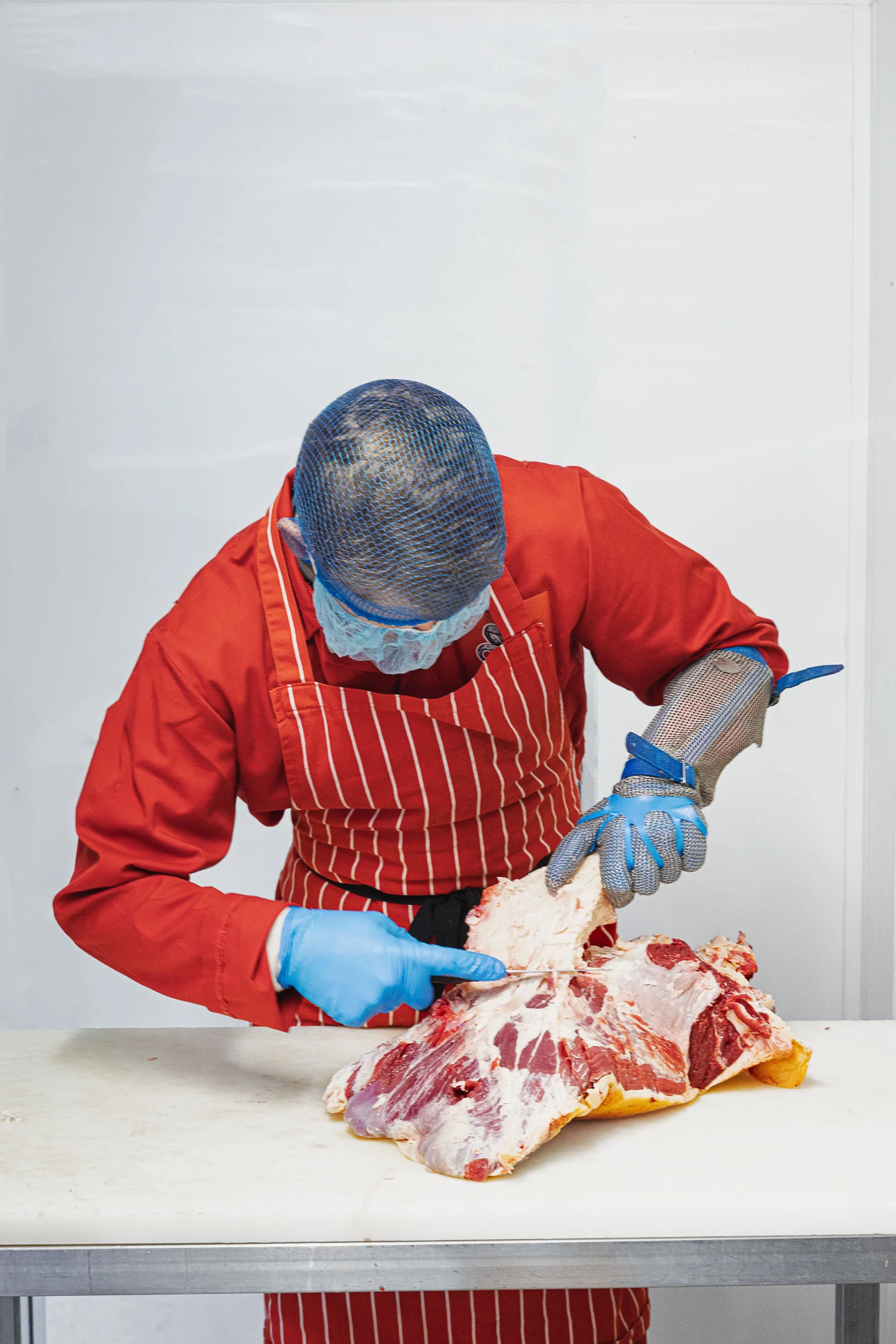 Documentary butchery portrait of a worker in protective clothing cutting and preparing raw meat on a clean production table, showing the skilled hand work and professional environment behind Sam Blunt bespoke butchery