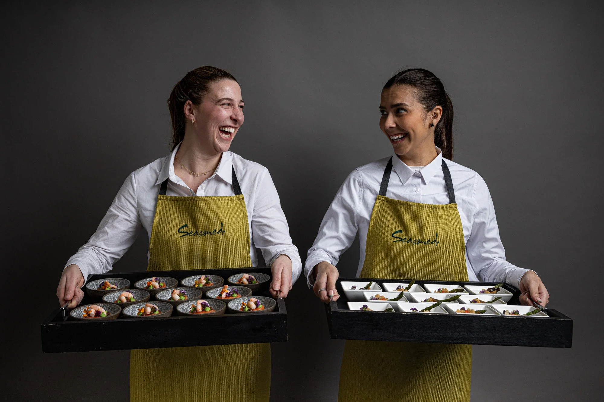 Commercial hospitality portrait of two Seasoned catering staff laughing while holding trays of individual dishes, using branded aprons and a clean dark background to present the team and food service together for promotional use.