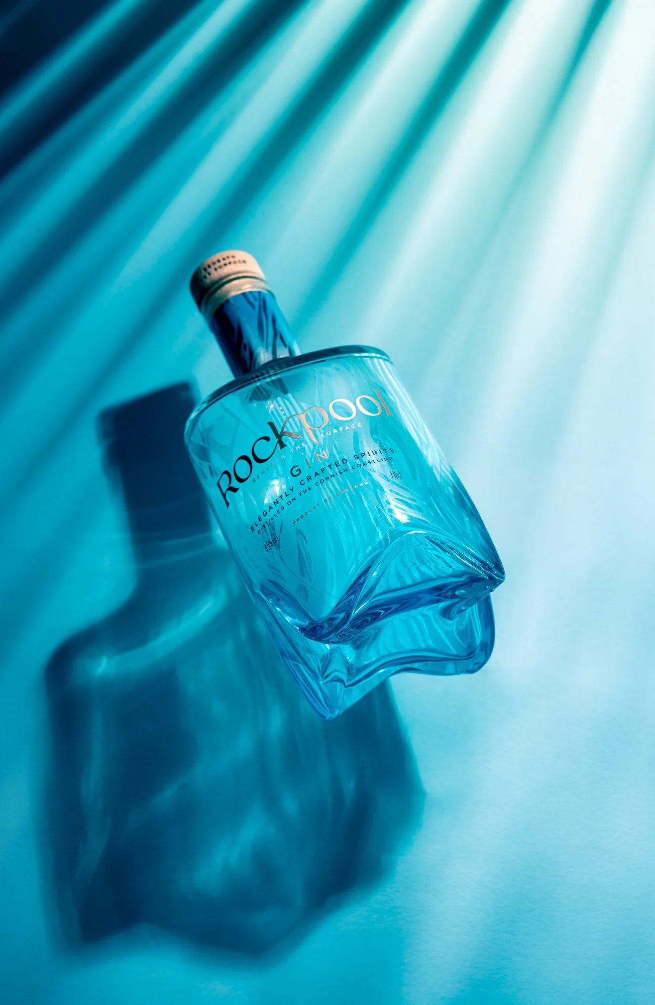 Commercial product photograph of a Rockpool Gin bottle lying on a vivid blue surface with strong directional shadows, using a tonal coastal palette to emphasise the clear glass, turquoise spirit and printed branding.